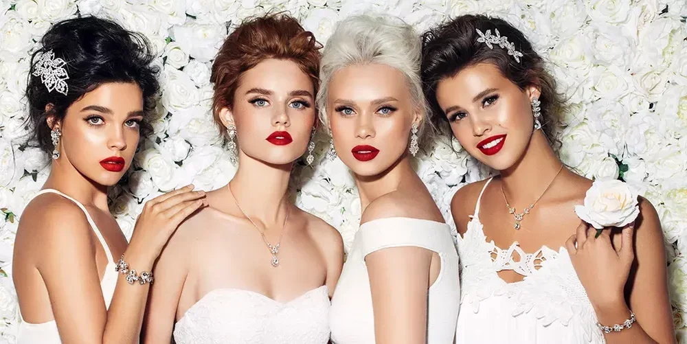 Four women dressed in white gowns with bridal accessories, posing in front of a white floral background.