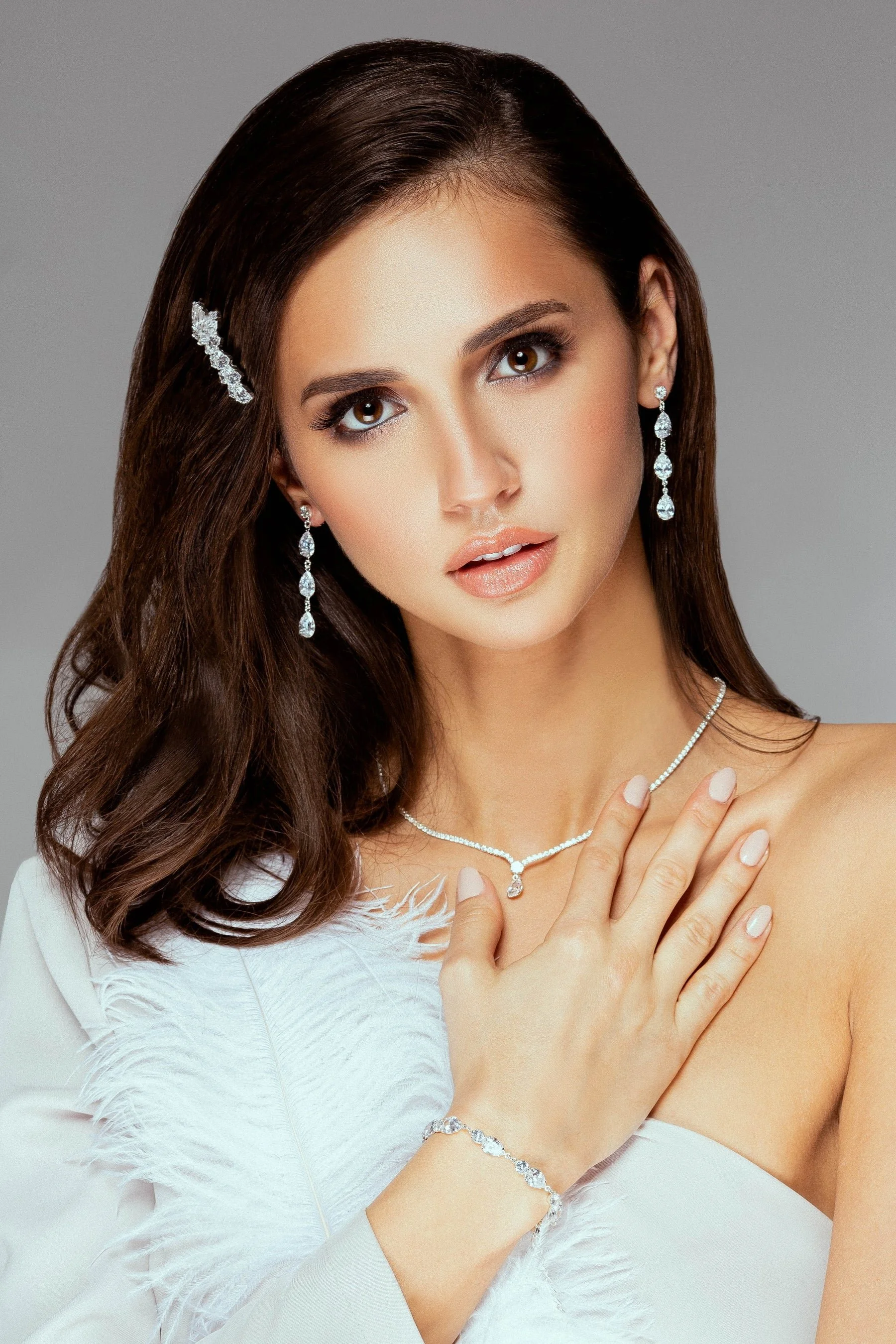 A woman with long brown hair wearing diamond jewelry, including earrings, a necklace, a bracelet, and a hair accessory, posing against a plain gray background.