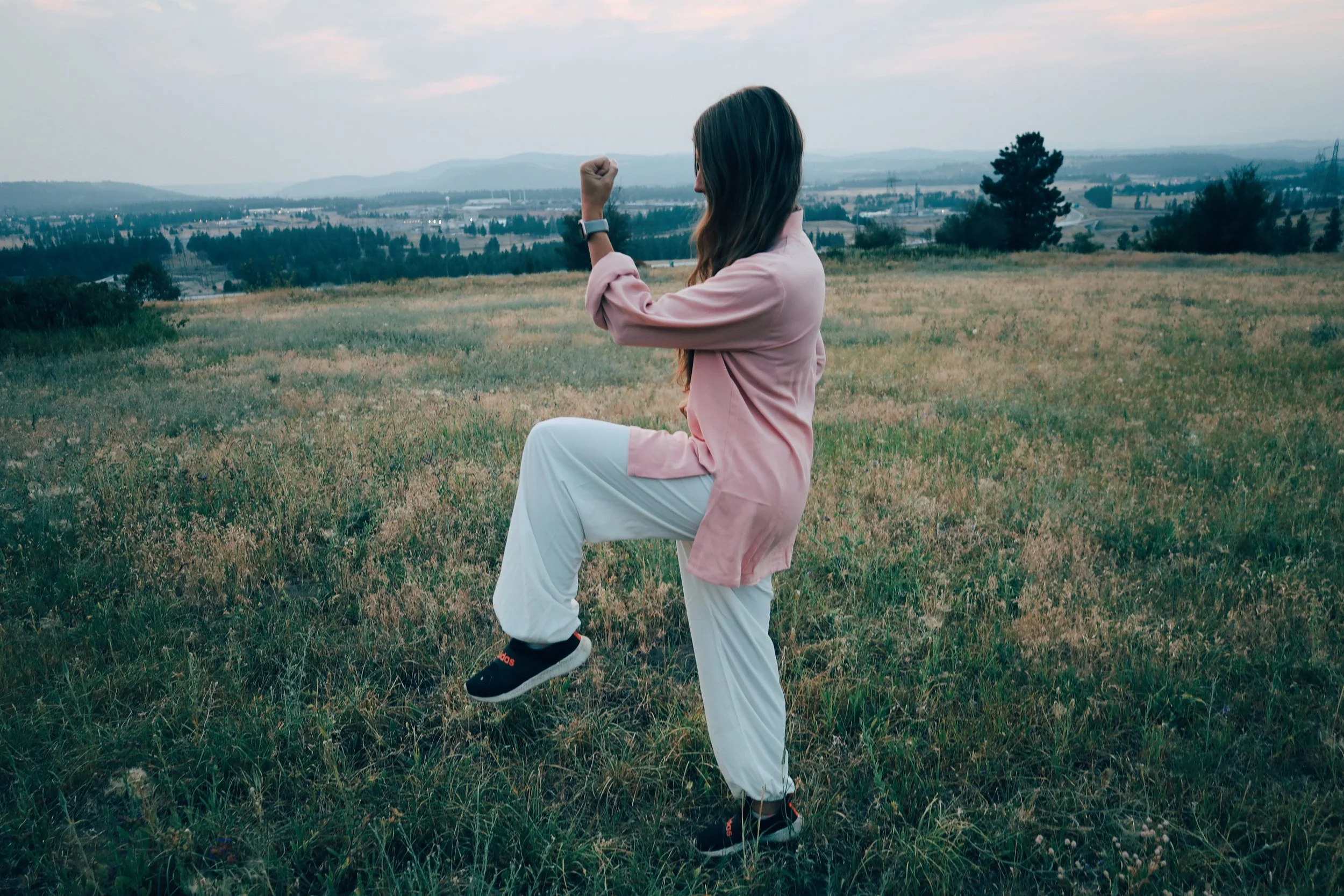 A woman with long brown hair wearing a pink jacket, white pants, and D Bal sneakers in a grass field, raising her right fist in the air with her left foot lifted, overlooking distant mountains and trees during sunset.