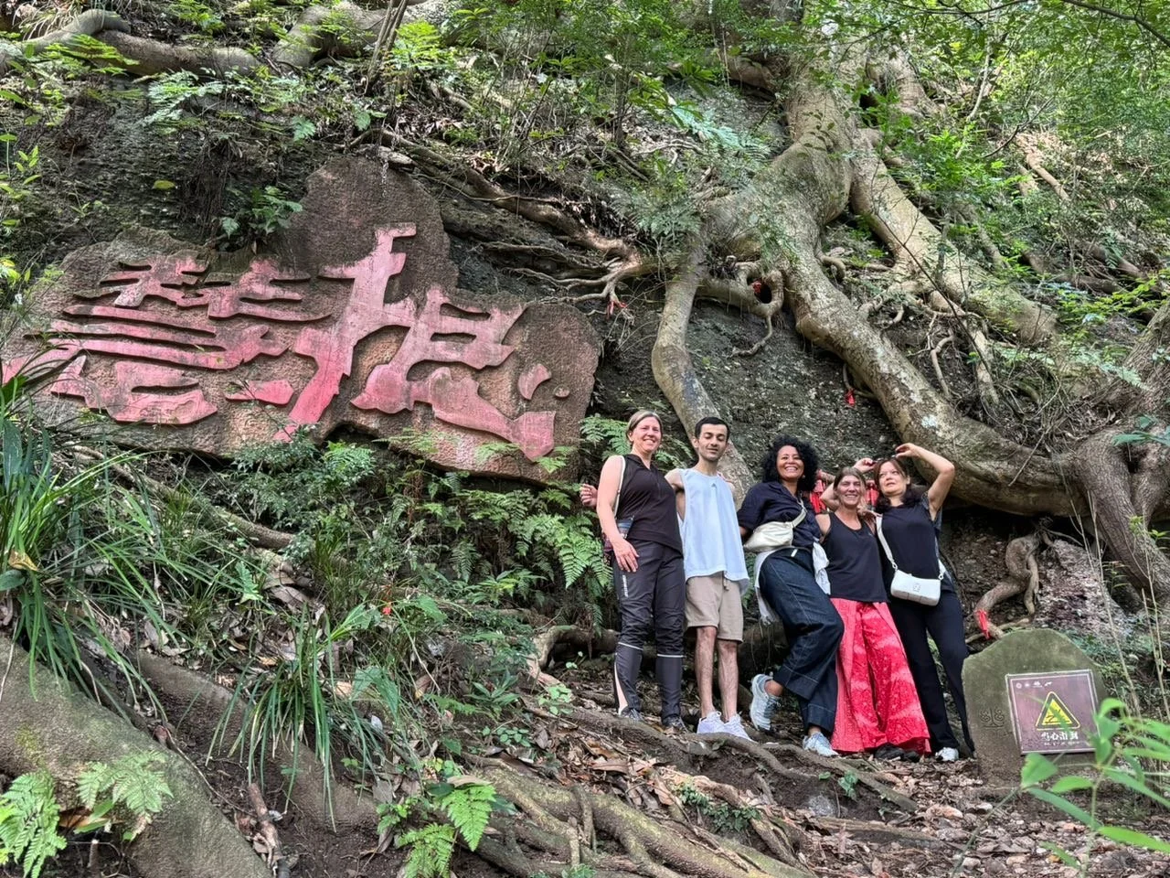 Five people standing on a trail in a forest, smiling at the camera, with large roots and green foliage around them. There is a large pink sign in the background and a small warning sign on a rock in the lower right corner.