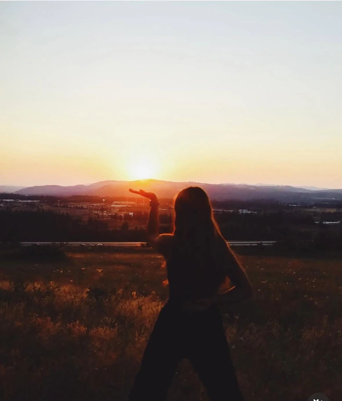 A person silhouette standing outdoors at sunset, with one arm raised as if holding the sun in their hand, overlooking a landscape with hills.