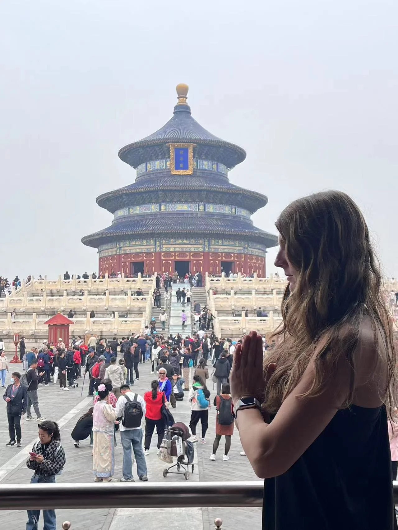 A woman with long wavy hair and a smartwatch on her wrist, standing in a praying posture with hands together, in front of the Temple of Heaven in Beijing, China, with many visitors in the background.