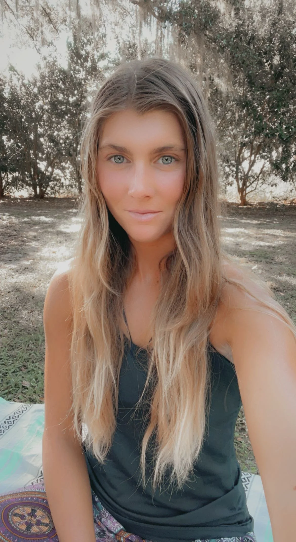 A woman with long wavy blonde hair and blue eyes sitting outdoors on a blanket, with trees and sunlight in the background.