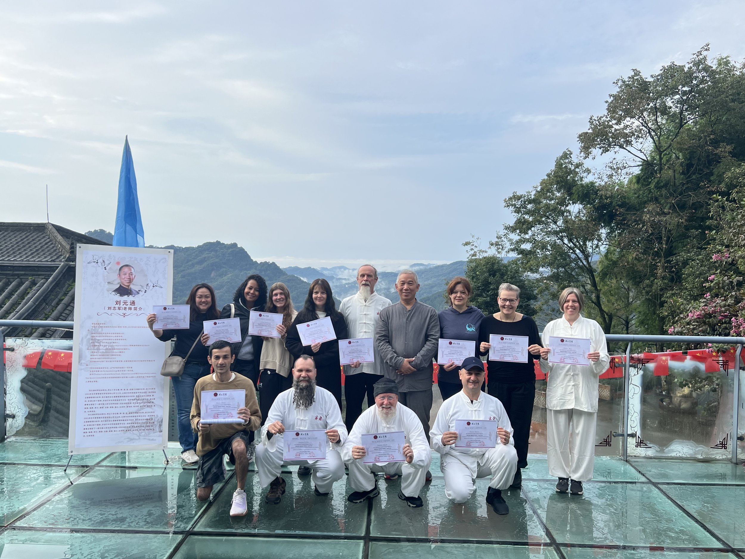 A group photo of people holding certificates outdoors on a cloudy day, with trees and mountains in the background.