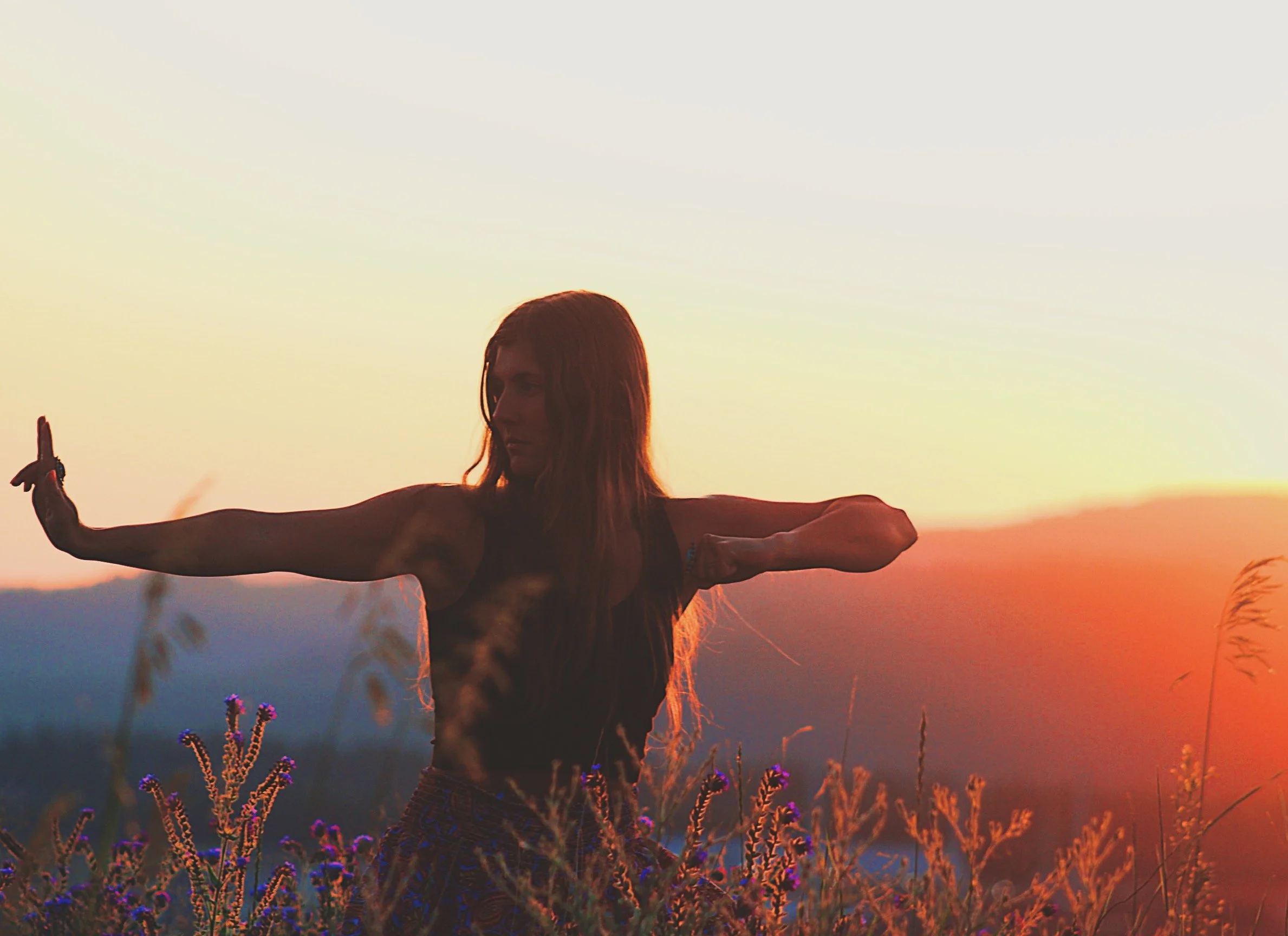 A woman practicing yoga outdoors at sunset, standing in a yoga pose with arms extended, surrounded by tall grass and wildflowers.