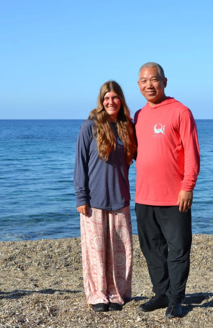 A smiling woman with long brown hair and a man with short gray hair standing together on a pebble beach near a body of water, with clear blue sky in the background.