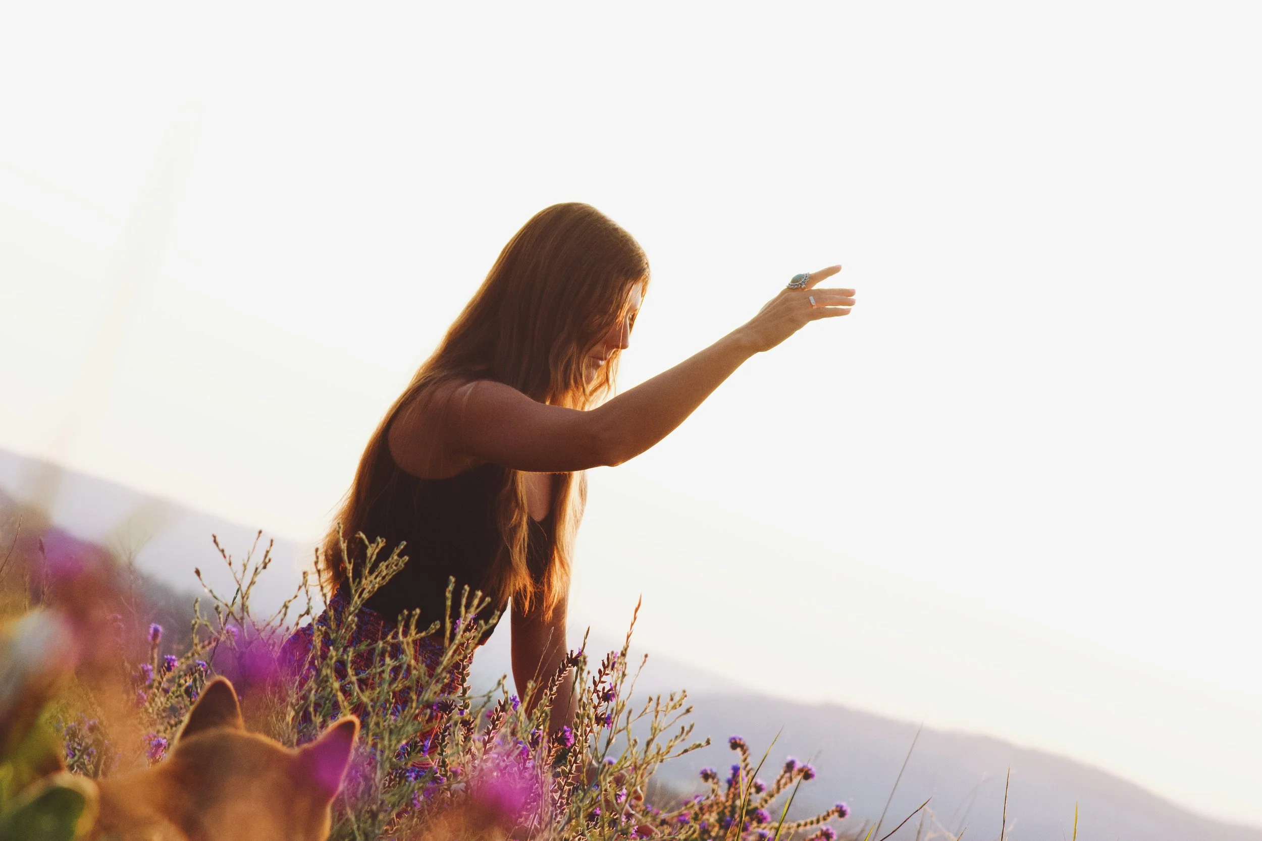 A woman with long red hair stands in a field of purple flowers, reaching out with her right arm, against a bright sky during sunset or sunrise.