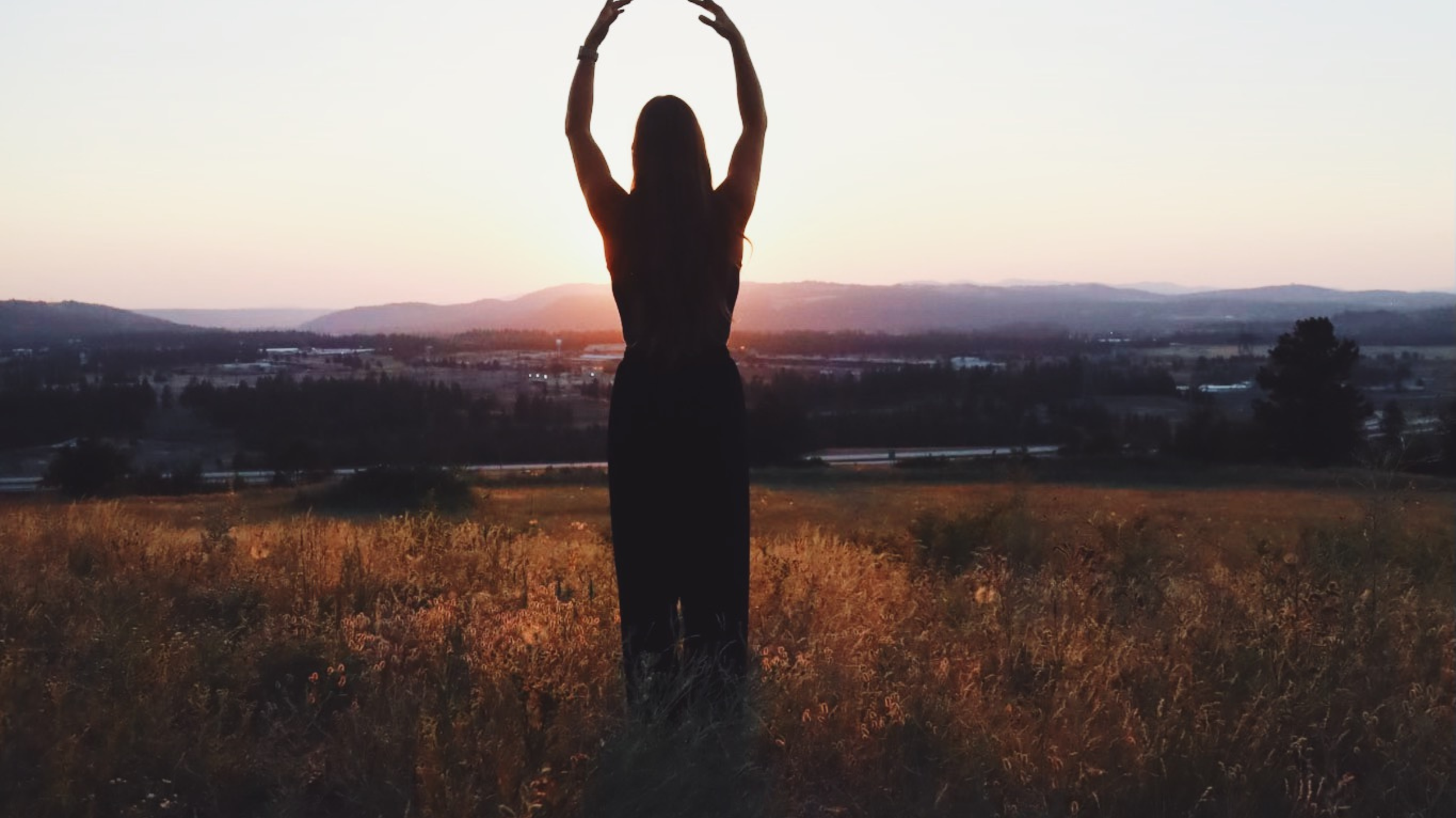 A woman stands outdoors during sunset, facing away with her arms raised and hands forming an arch over her head, overlooking a landscape of fields and distant hills.