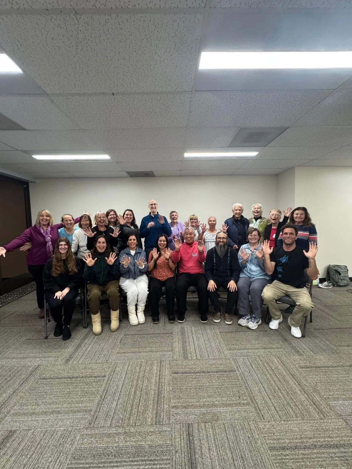 A group of people posing for a photo in an indoor room, many waving and smiling at the camera.