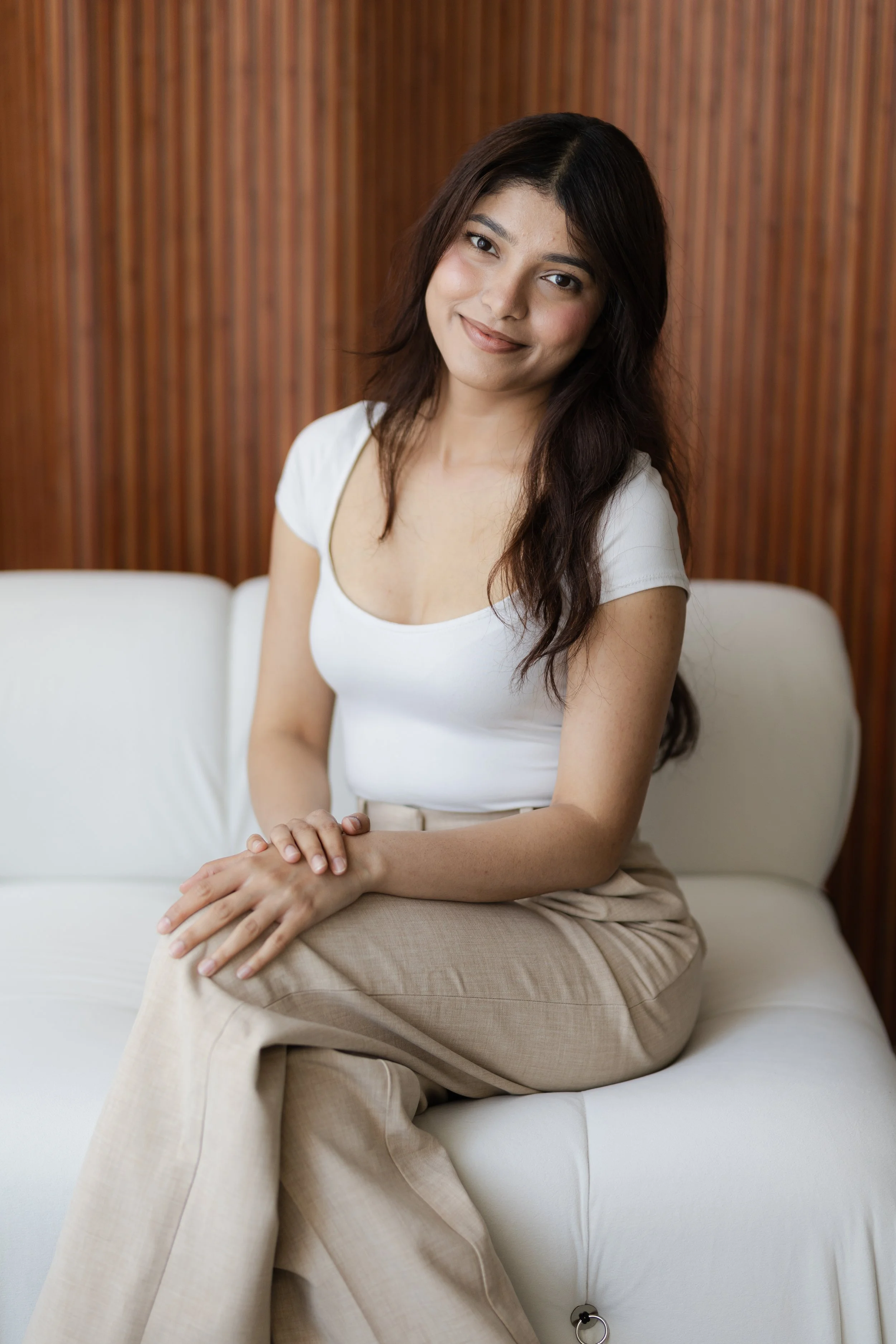 A young woman with dark hair and a white top sitting on a white sofa with her hands on her lap, in front of a wooden paneled wall, smiling at the camera.