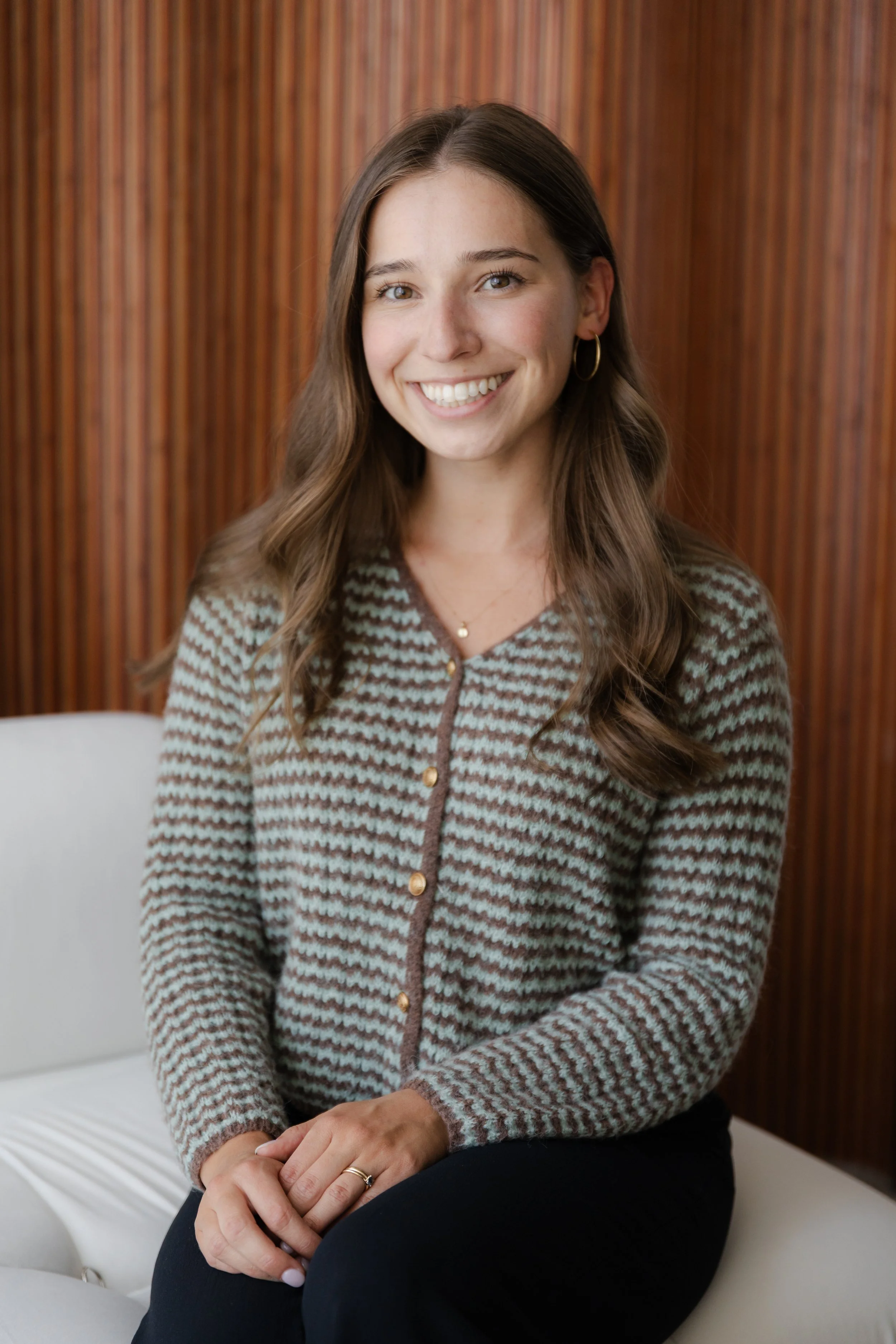 A woman with long brown hair smiling, sitting on a white couch with a wooden striped wall in the background. She wears a patterned sweater, gold hoop earrings, and a gold necklace.