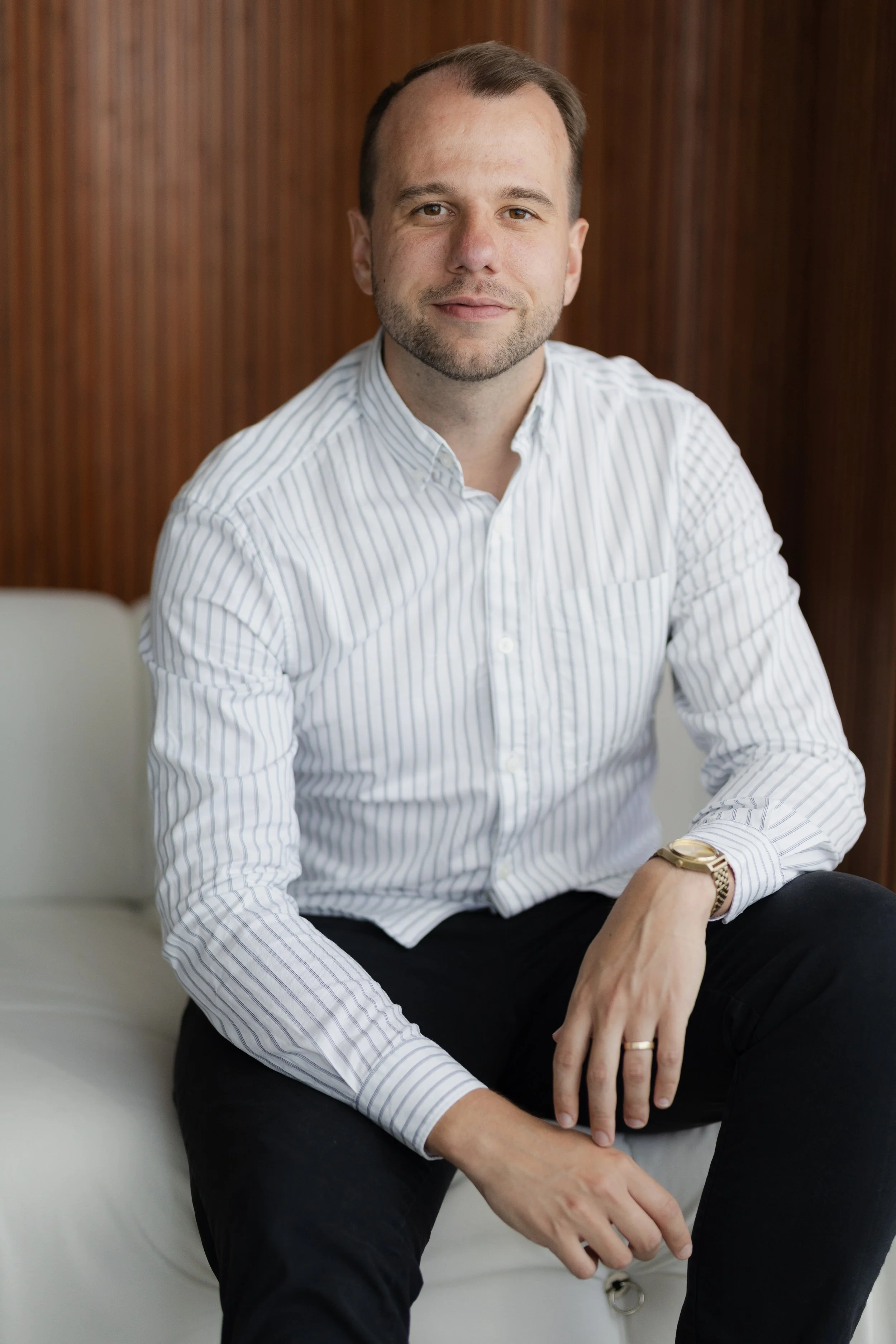 A man with short brown hair, light skin, and a beard, sitting indoors against a wooden wall, wearing a white striped dress shirt, a gold watch, and rings, with his right hand resting on his thigh and his left arm resting on the armrest.