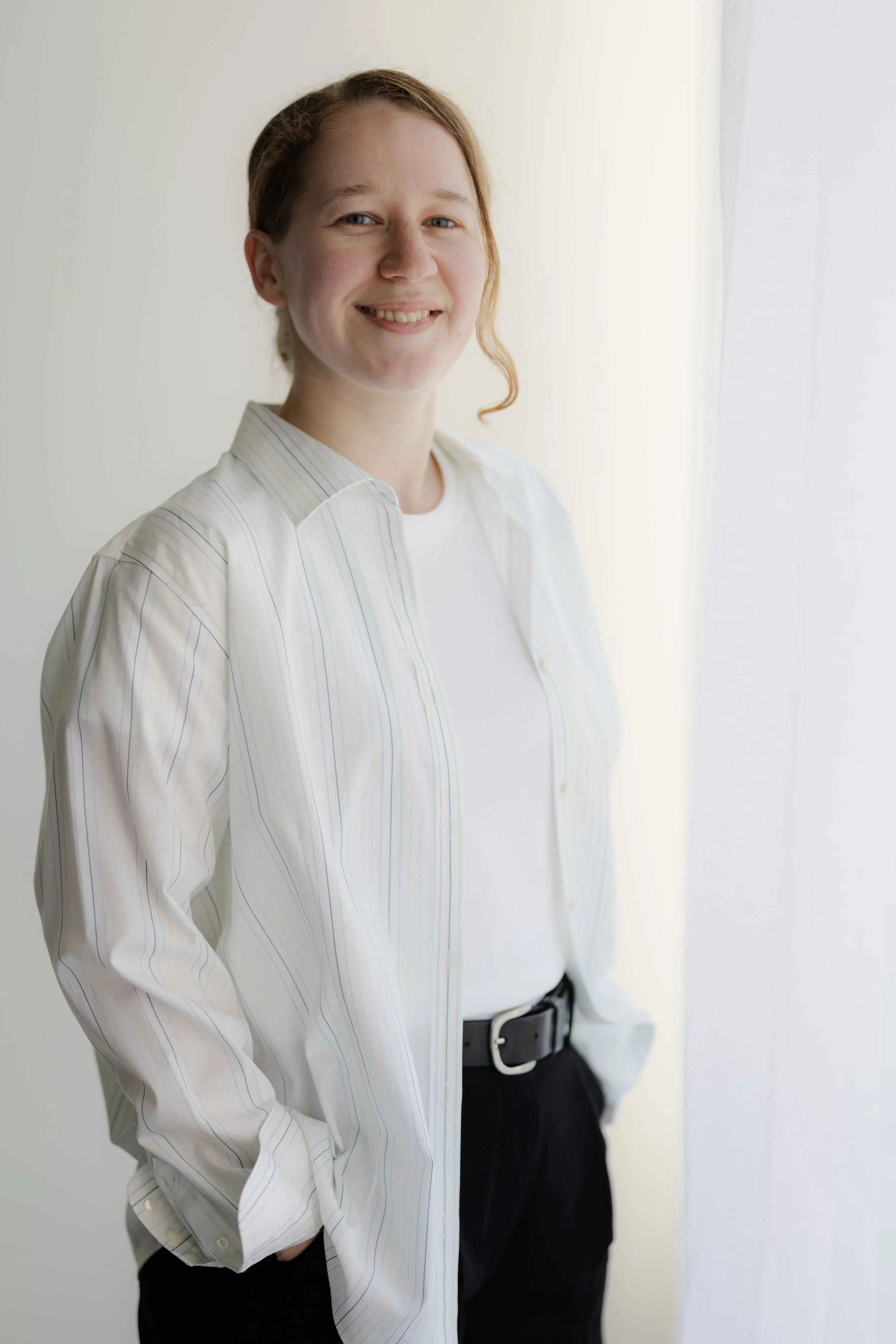 Young woman with light skin, red hair, and blue eyes, wearing a white shirt with thin blue stripes and dark pants, standing near a window with sheer curtains in natural light.