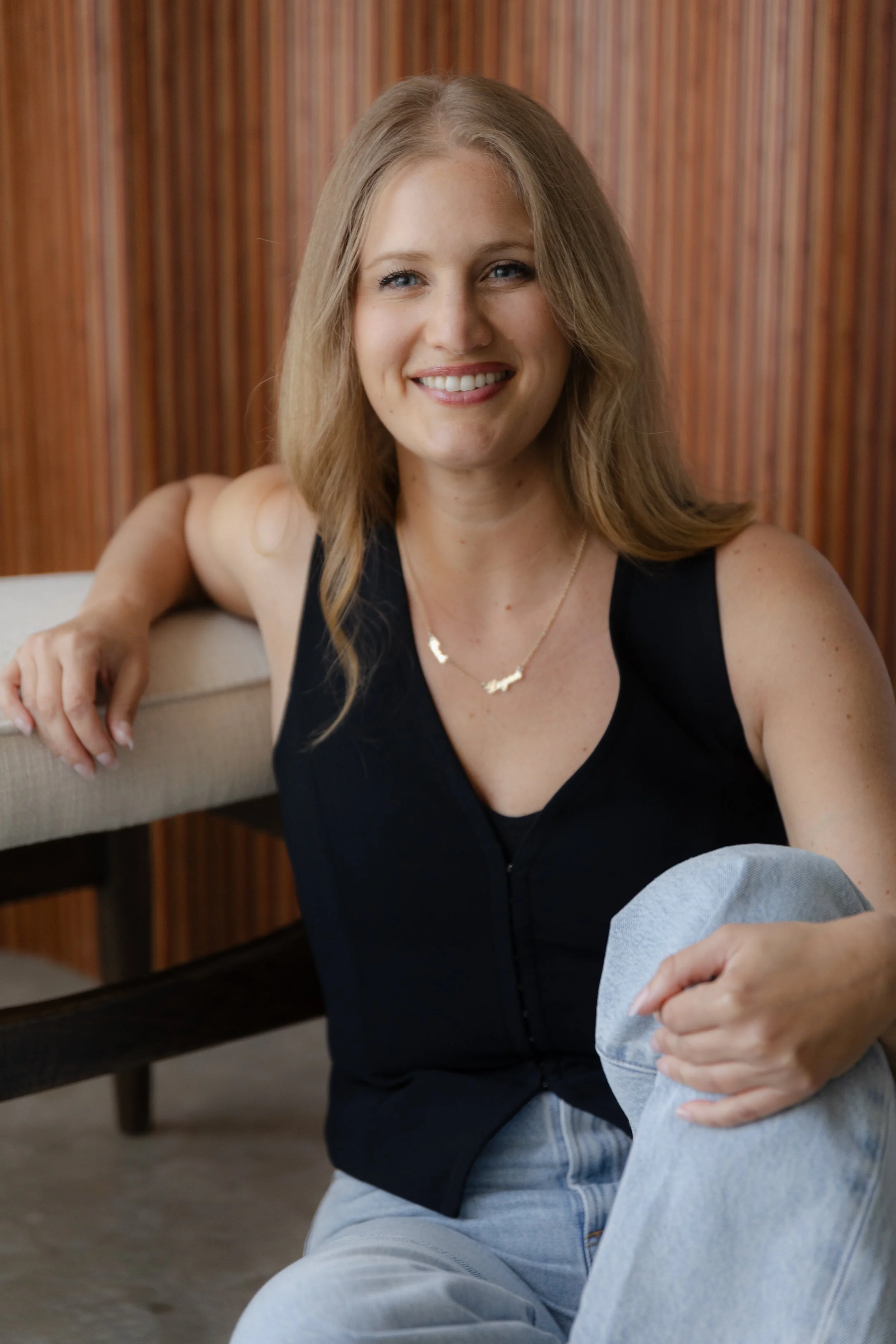 A woman with long blonde hair, wearing a black top and a gold necklace, smiling while sitting on a beige couch with a wooden wall background.