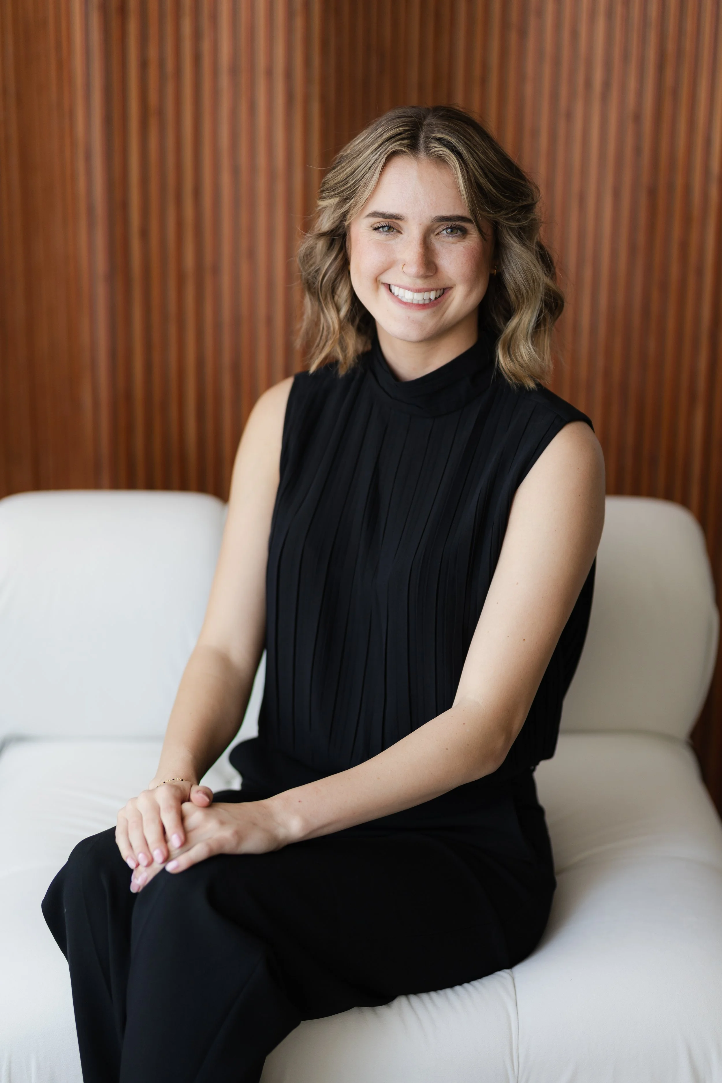 A young woman with blonde, wavy hair, smiling, wearing a sleeveless black top, sitting on a white sofa against a wooden paneled wall.