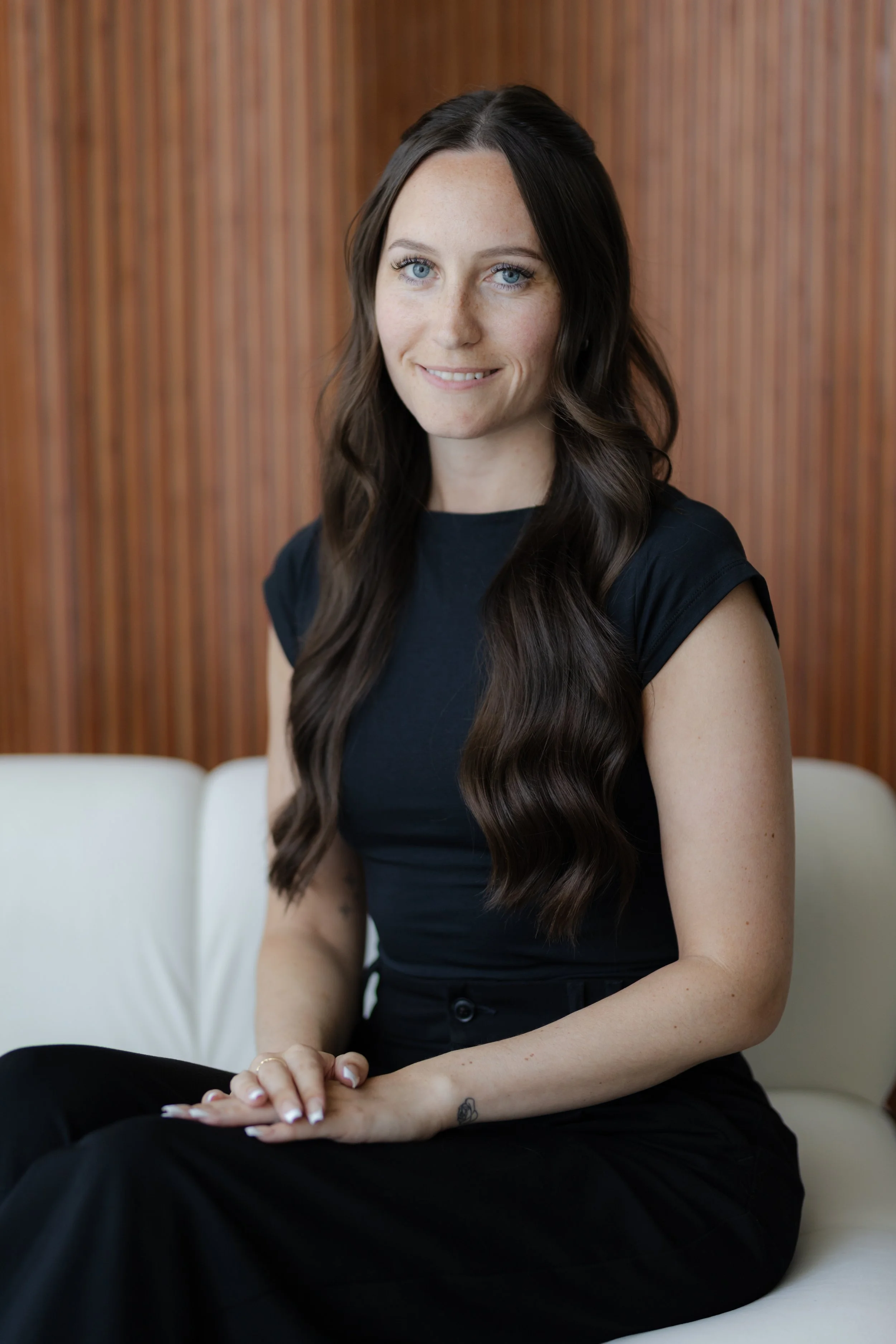 A woman with long wavy dark hair, blue eyes, and light freckles is sitting on a white couch with a wooden panel background, smiling at the camera. She is wearing a black short-sleeved top.