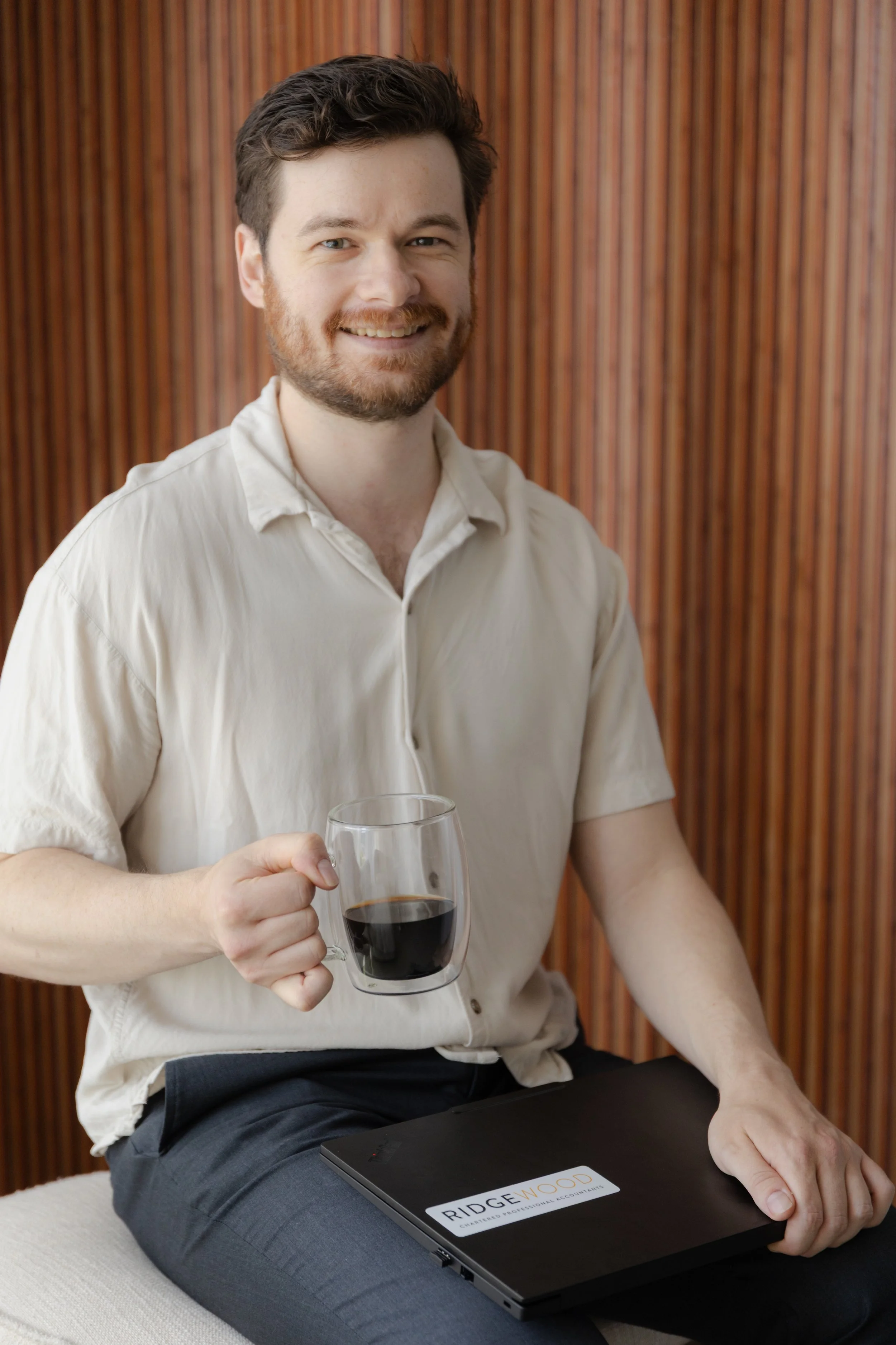 A man with brown hair, a beard, and a smile, sitting in front of a wooden paneled wall, holding a glass of red wine in his right hand and resting his left hand on a black Ridgewood Chartered Professional Accountants folder.
