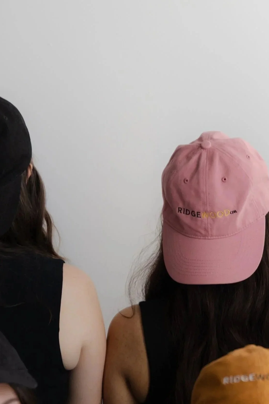 Two women wearing black tank tops and hats, one with a pink cap reading 'RIDGEWOOD', sitting against a plain white wall.