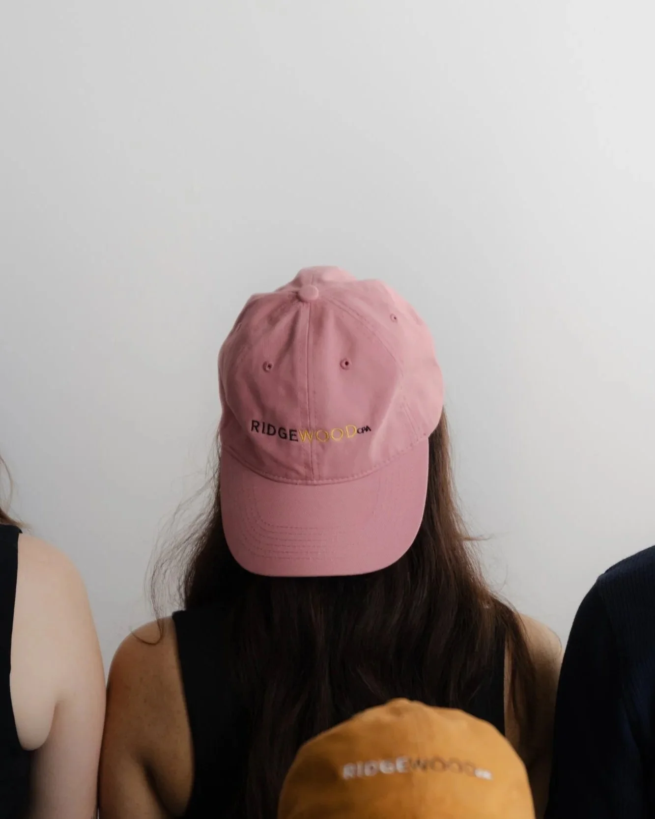Person wearing a pink cap with the words "RIDGEWOOD" embroidered on it, partially obscuring their face. They have long dark hair and are wearing a black tank top. Two other people are partially visible on either side of the person, and the background is a plain white wall.