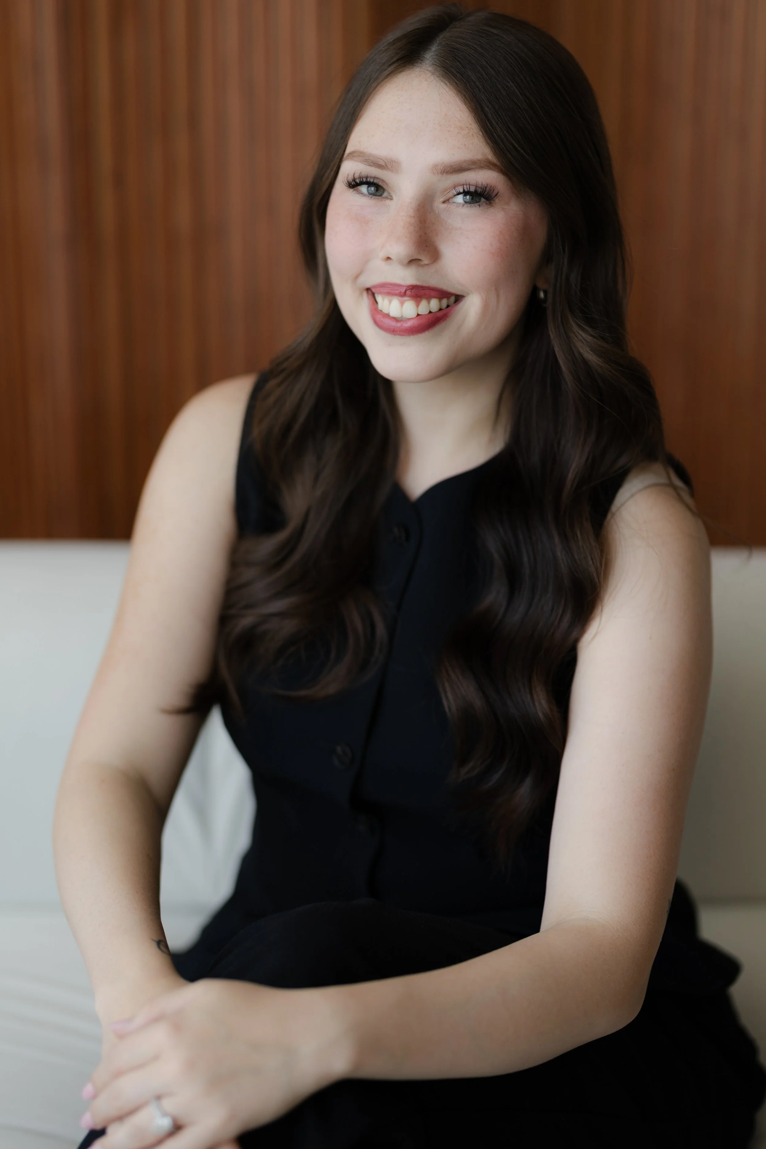 A young woman with long wavy brown hair and blue eyes, smiling with white teeth, sitting indoors with a wooden wall background, wearing a black sleeveless top.