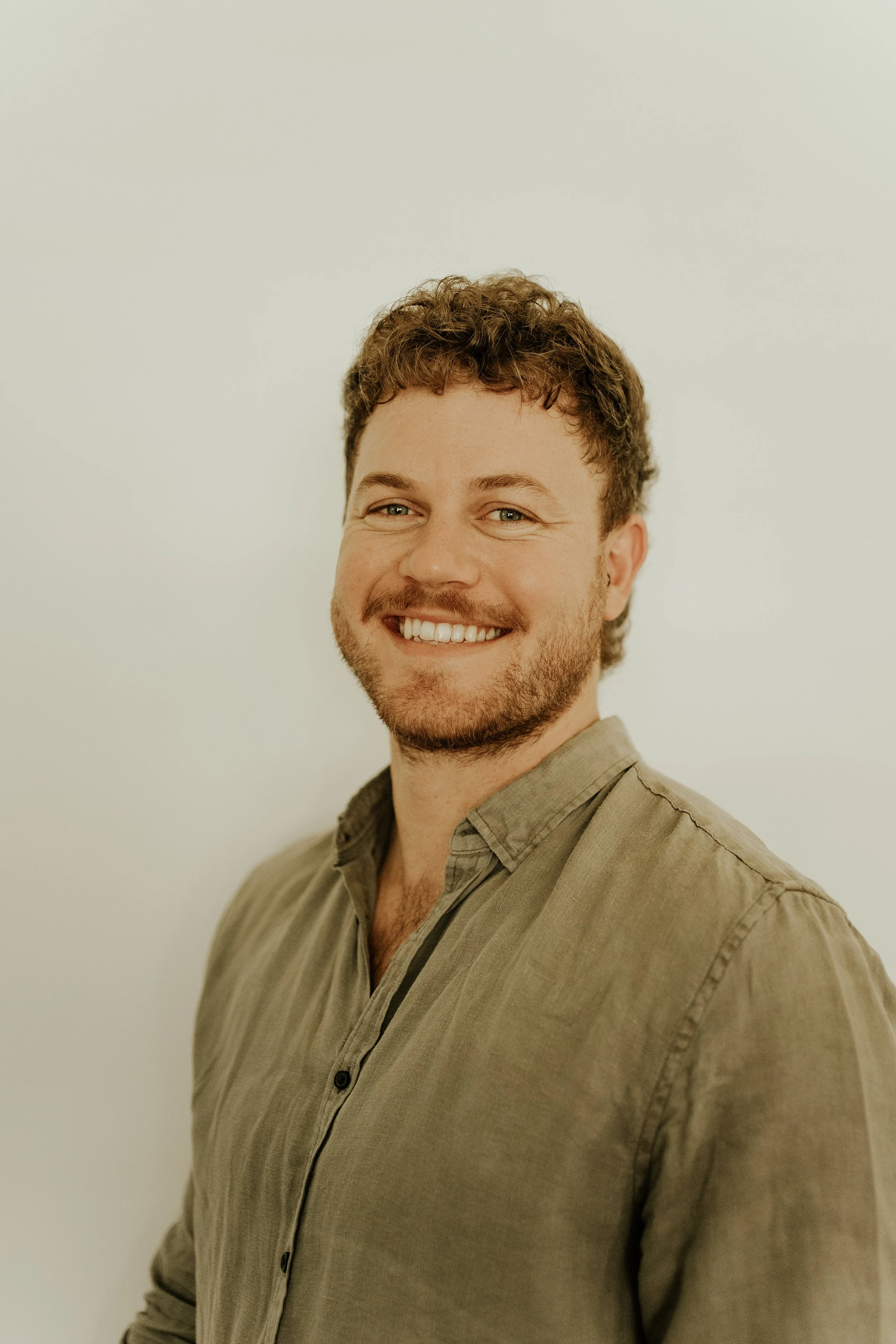 Smiling man with curly brown hair, beard, blue eyes, wearing a beige button-up shirt, standing against a plain white wall.