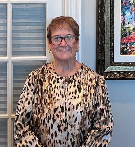 A smiling elderly woman with short reddish-brown hair and glasses, wearing a leopard print blouse, standing indoors near a window and a framed colorful abstract painting.
