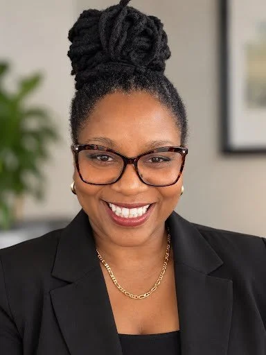 A woman with dark braided hair styled in a high bun, wearing glasses, a black blazer, gold earrings, and a gold chain necklace, smiling in an indoor setting.