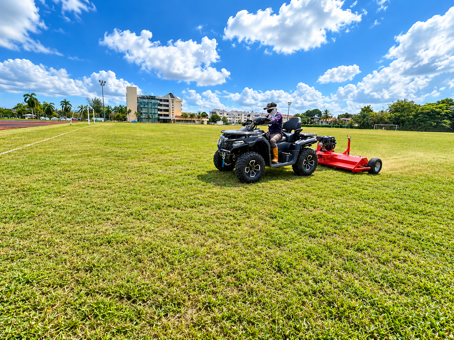 A person operating a black ATV on a grassy field with a red trail mower attached, under a partly cloudy blue sky, with modern buildings and palm trees in the background.