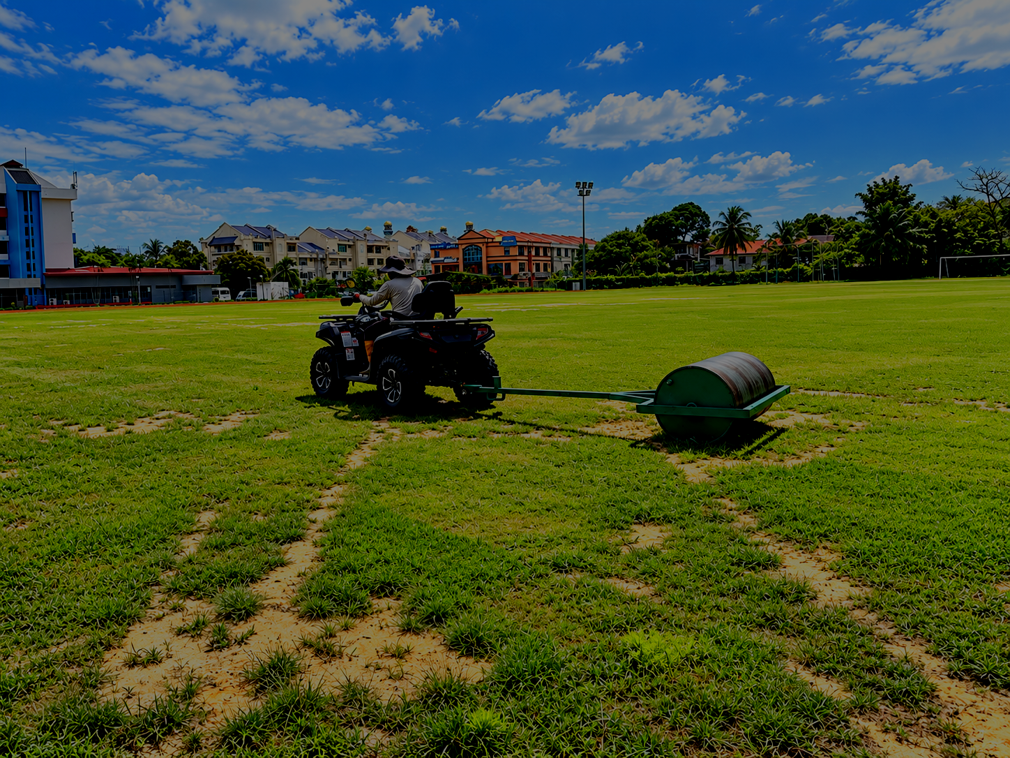 A person riding an all-terrain vehicle (ATV) pulling a roller to water or fertilize a grassy field on a sunny day with partly cloudy skies, with residential buildings and trees in the background.