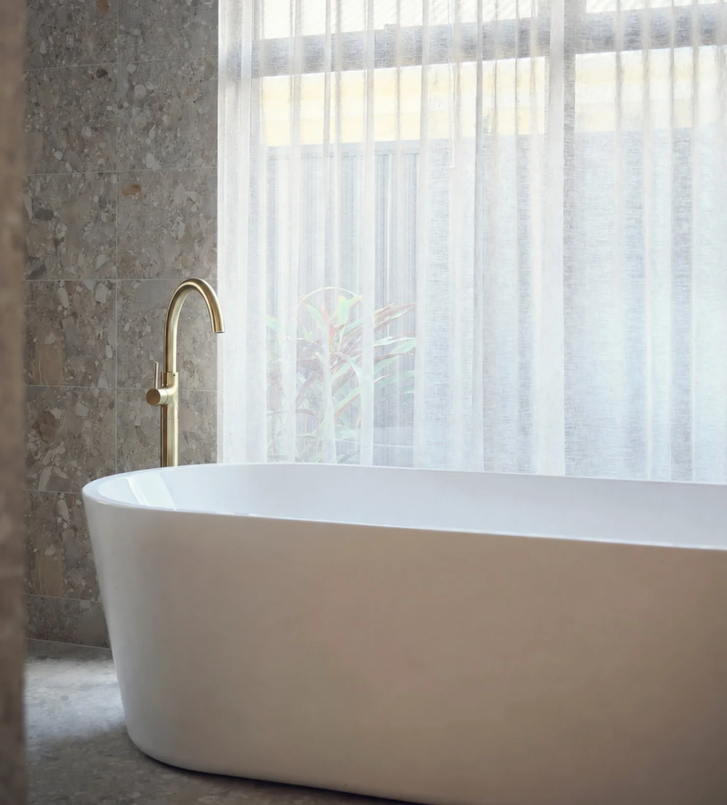 Curved freestanding bath beside sheer curtains, with a brass floor-mounted tap and textured stone wall
