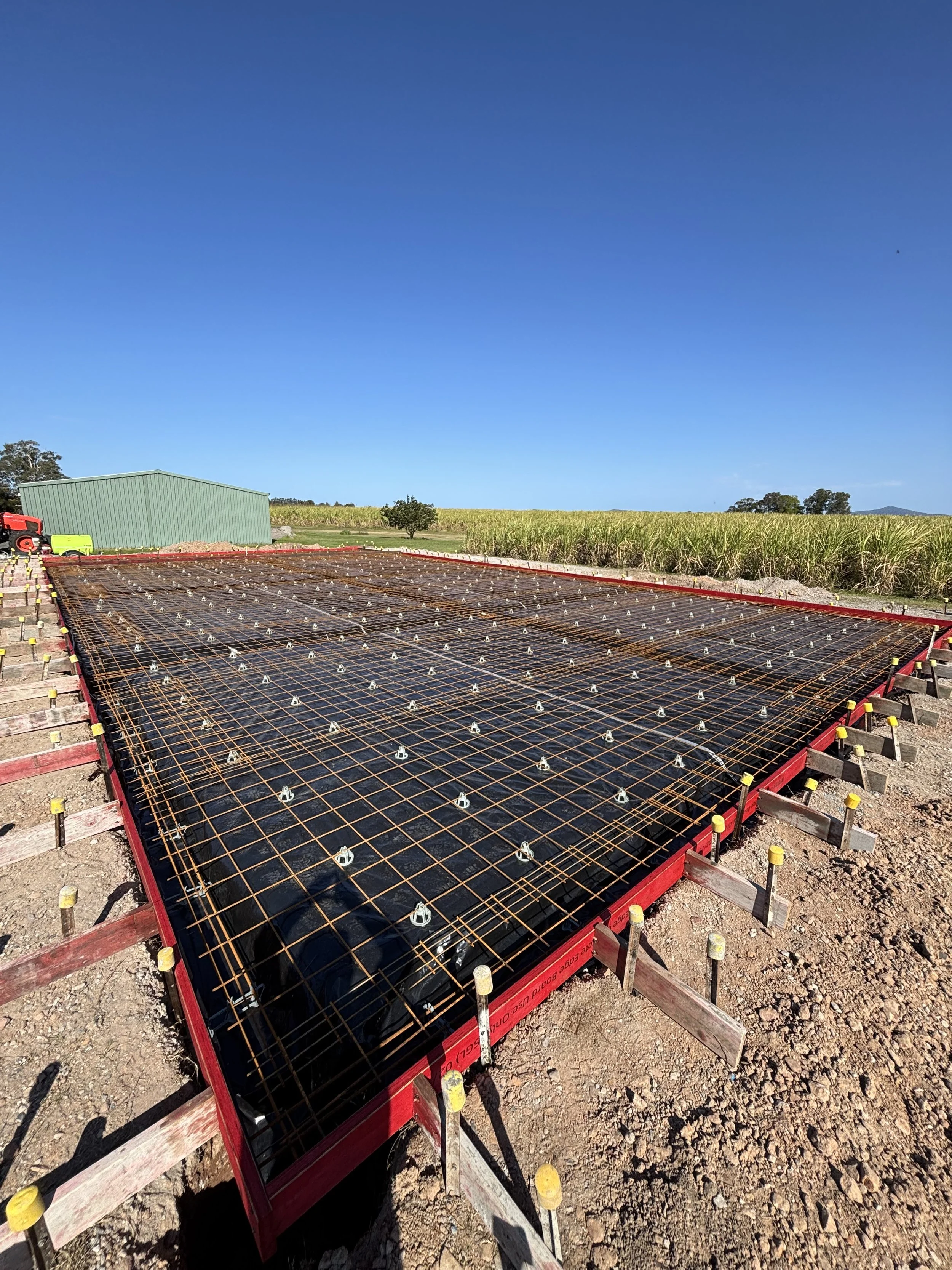 Construction site of a shed slab with reinforced concrete foundation in progress, metal rebar grid, and a green barn in the background, under a clear blue sky in Yamba.