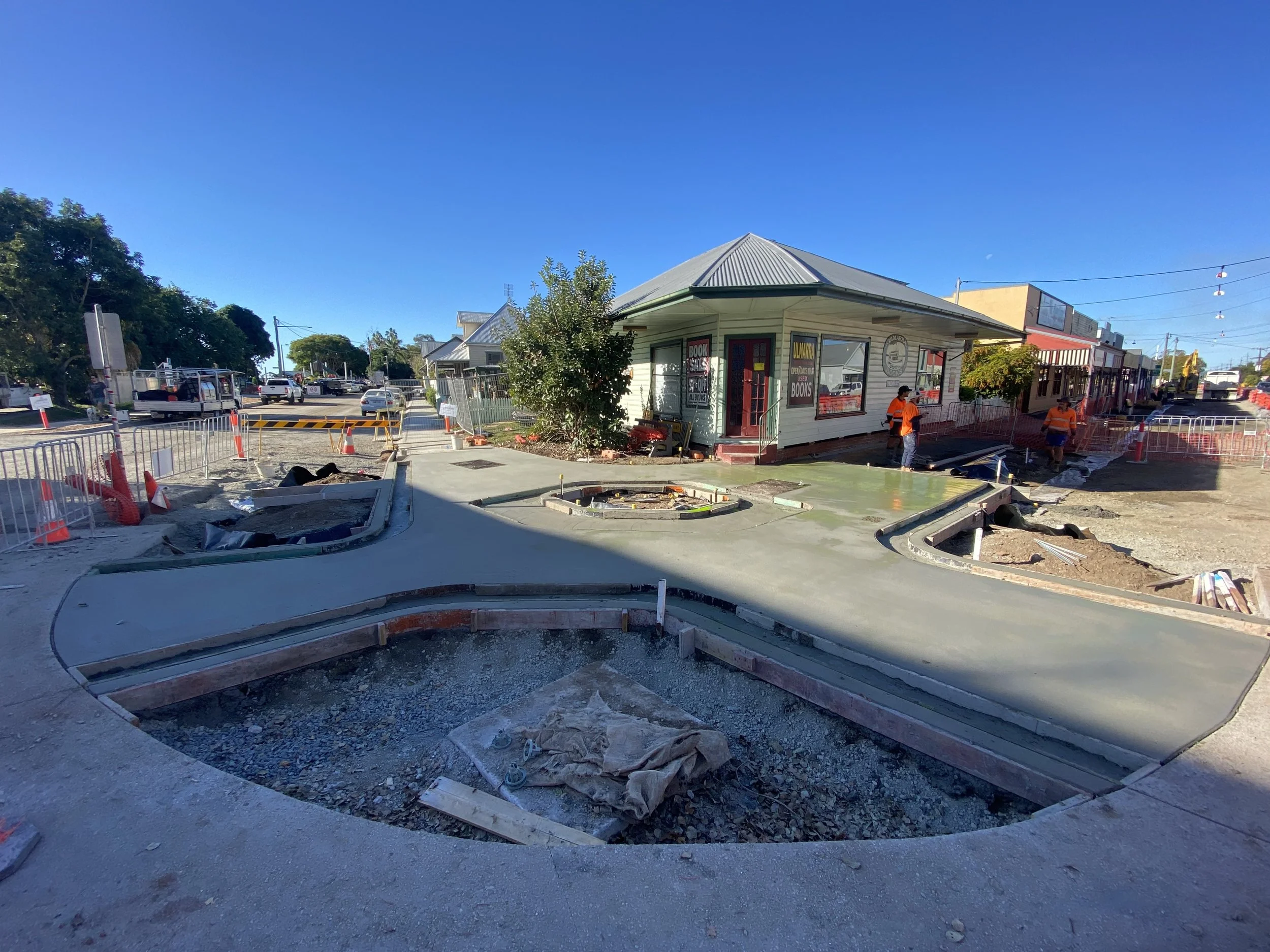 Street view showing civil construction and concrete work on a footpath with a small building, workers, barriers, and vehicles, for the Clarence Valley council in Ulmarra.
