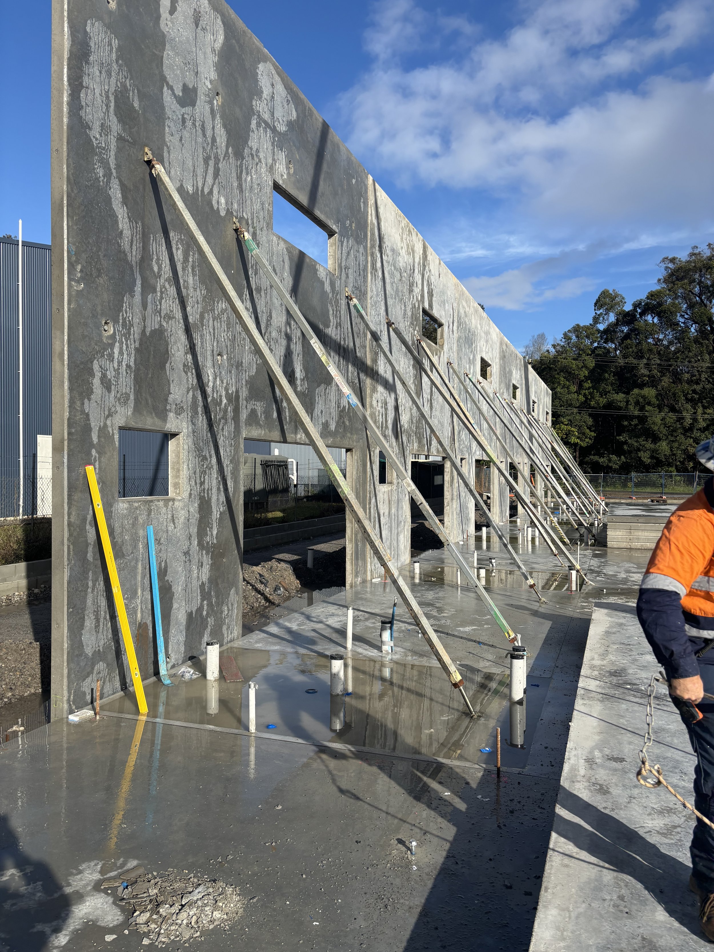 A commercial tilt panel construction site in Grafton with a large concrete wall supported by diagonal braces, small windows in the wall, finished concrete on the ground, and some construction tools and equipment visible.