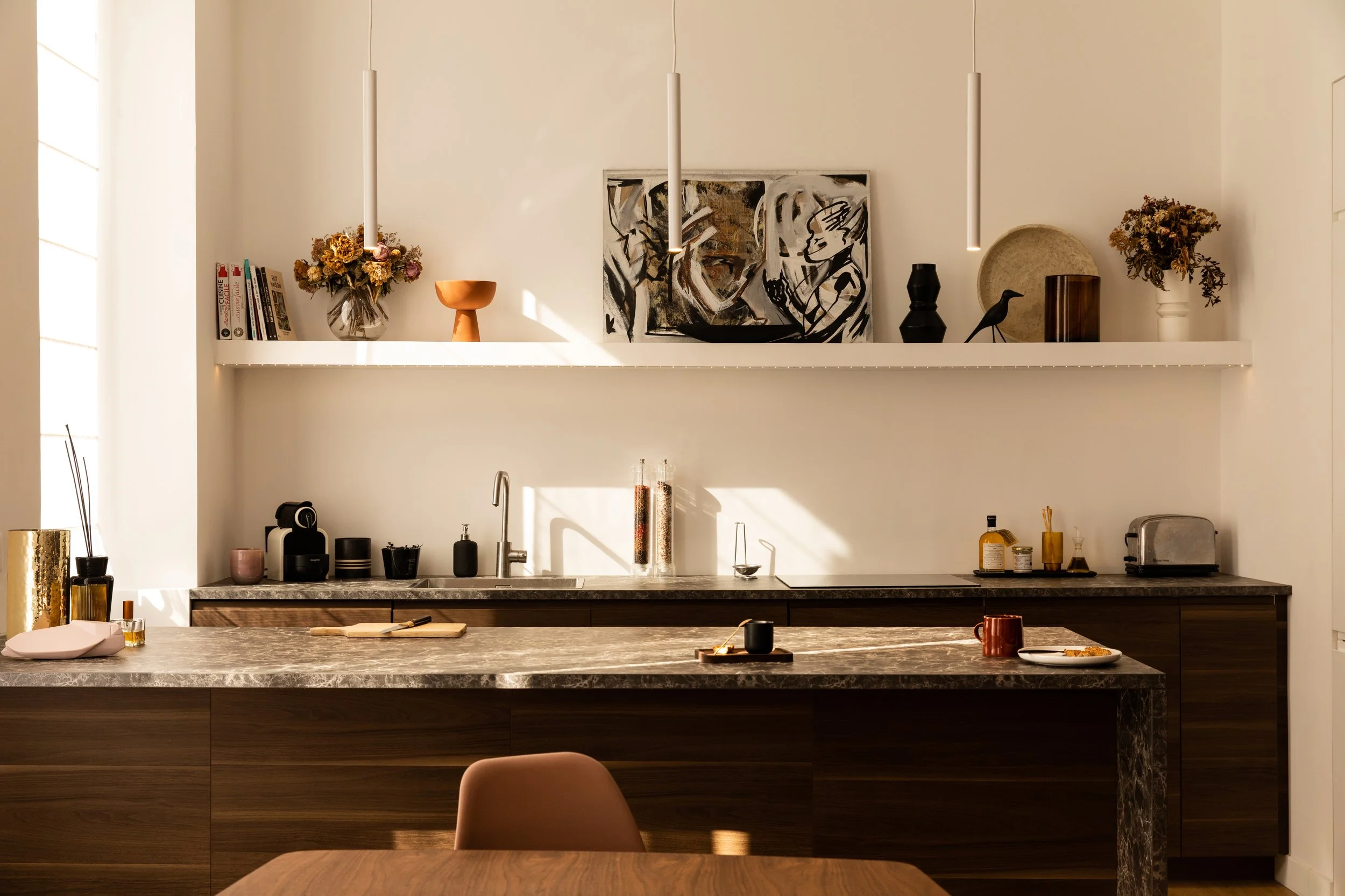 Cuisine moderne avec une étagère décorative contenant des livres, des vases avec des fleurs séchées, une peinture abstraite, un bol en terre cuite, une sculpture de cire noire, une vaisselle en céramique blanche, un café et quelques outils de cuisine sur le plan de travail en marbre.