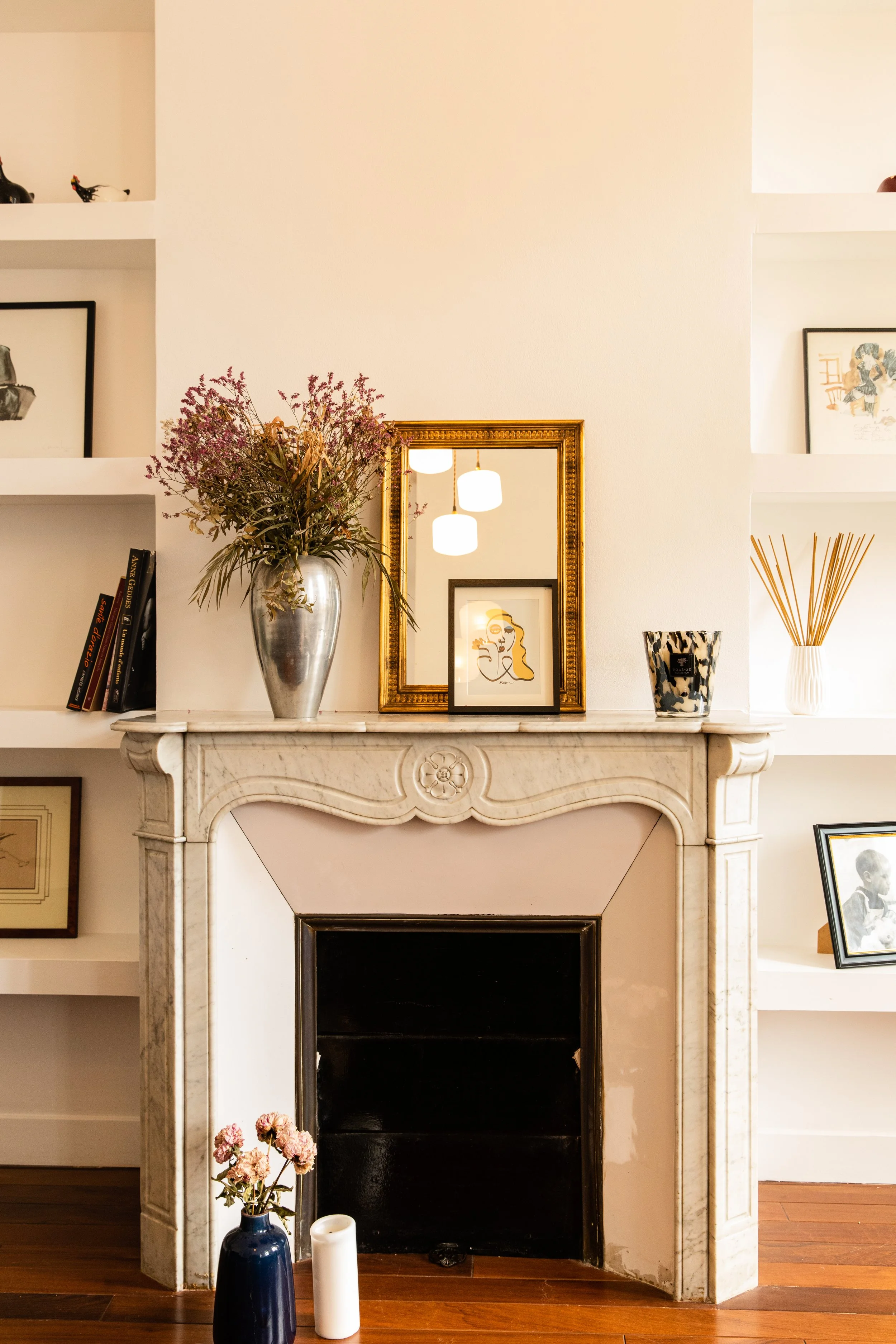 Une cheminée en marbre blanc dans un salon, décorée avec un vase contenant des fleurs, un miroir doré, et des cadres au mur, avec des bougies en premier plan.