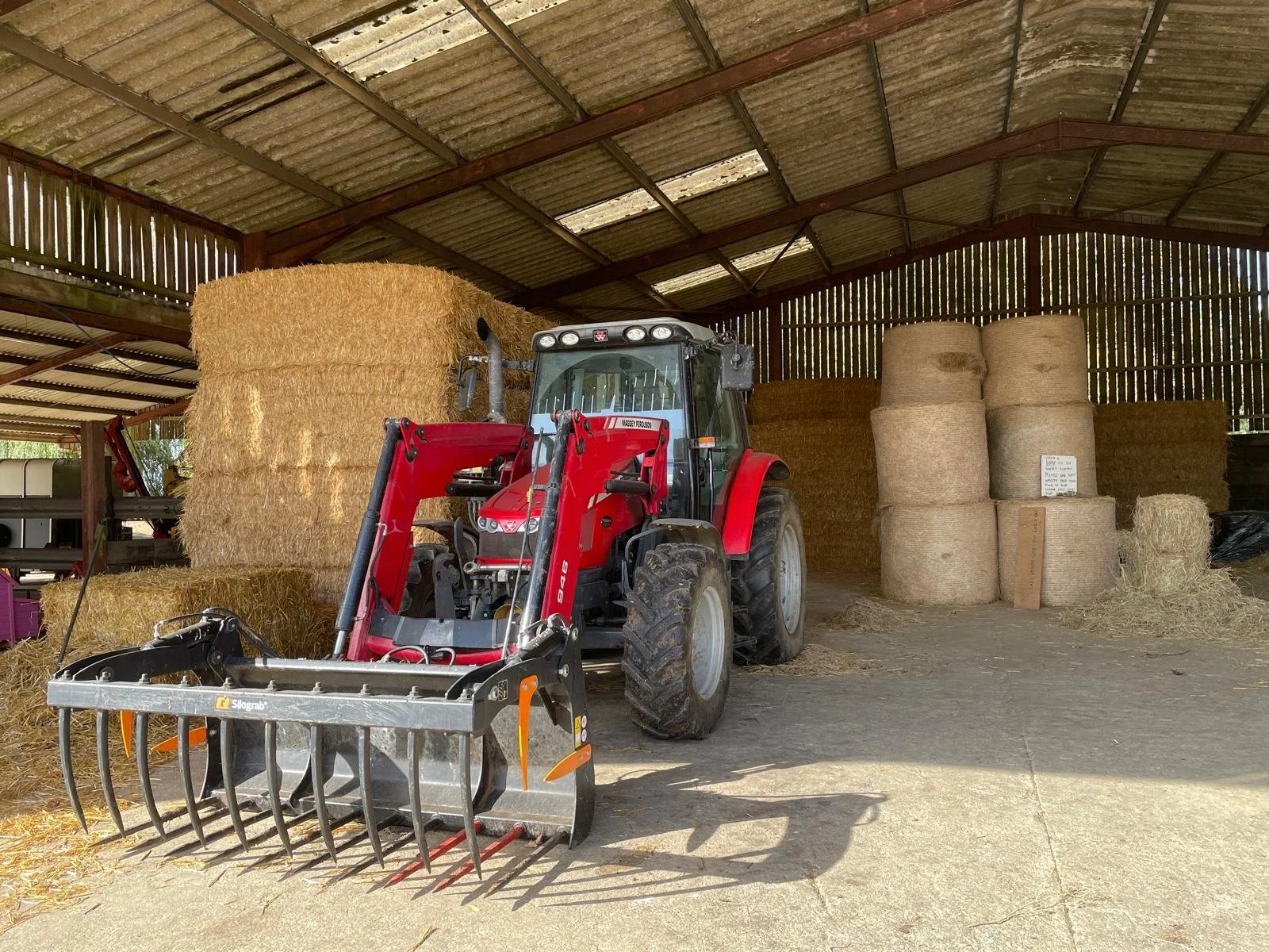 Inside a barn with a red tractor and large hay bales, some wrapped and some loose, under a metal roof with wooden panels.