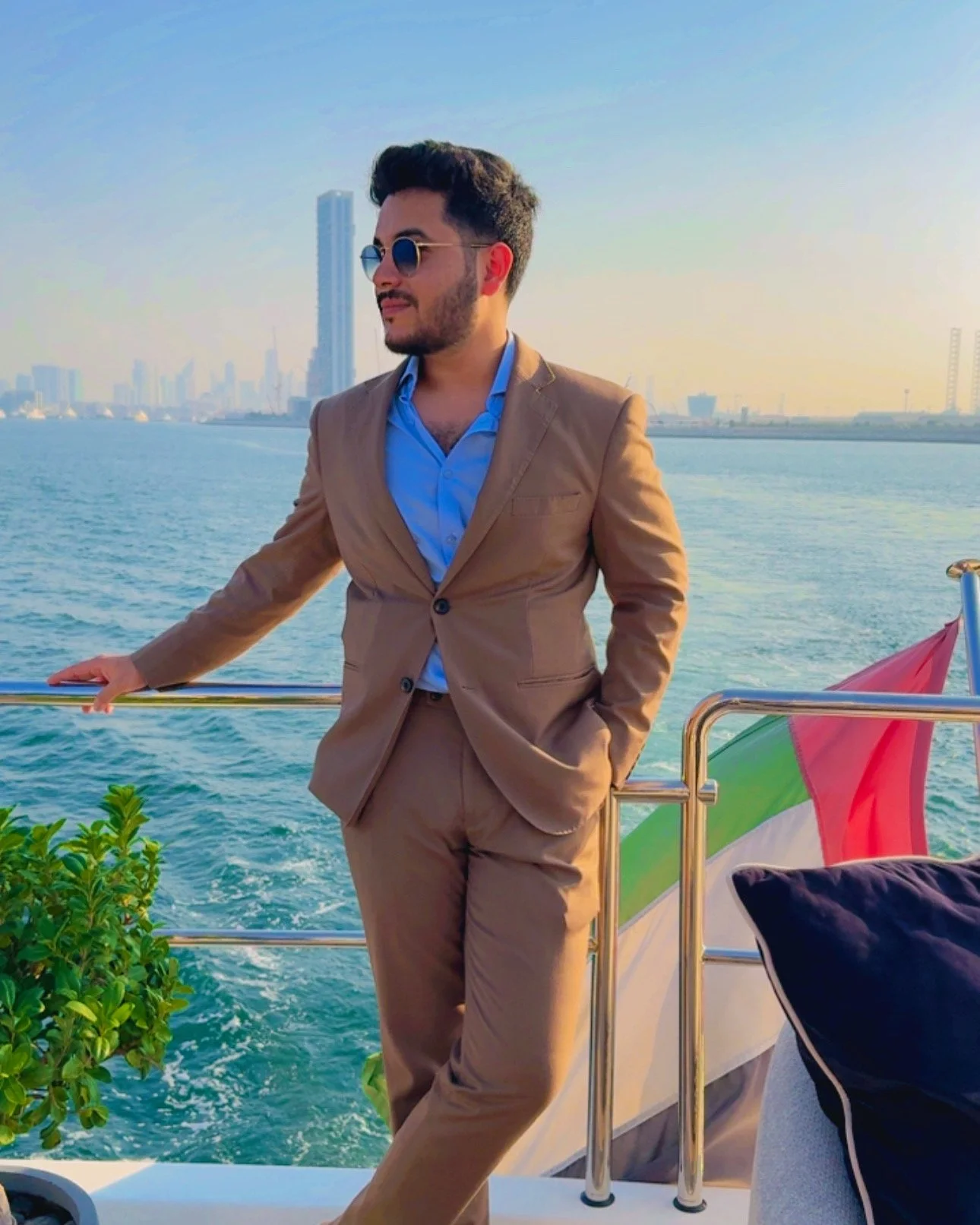A man in a brown suit and blue shirt wearing sunglasses, standing on a boat with a city skyline in the background, and an Italian flag on the boat.