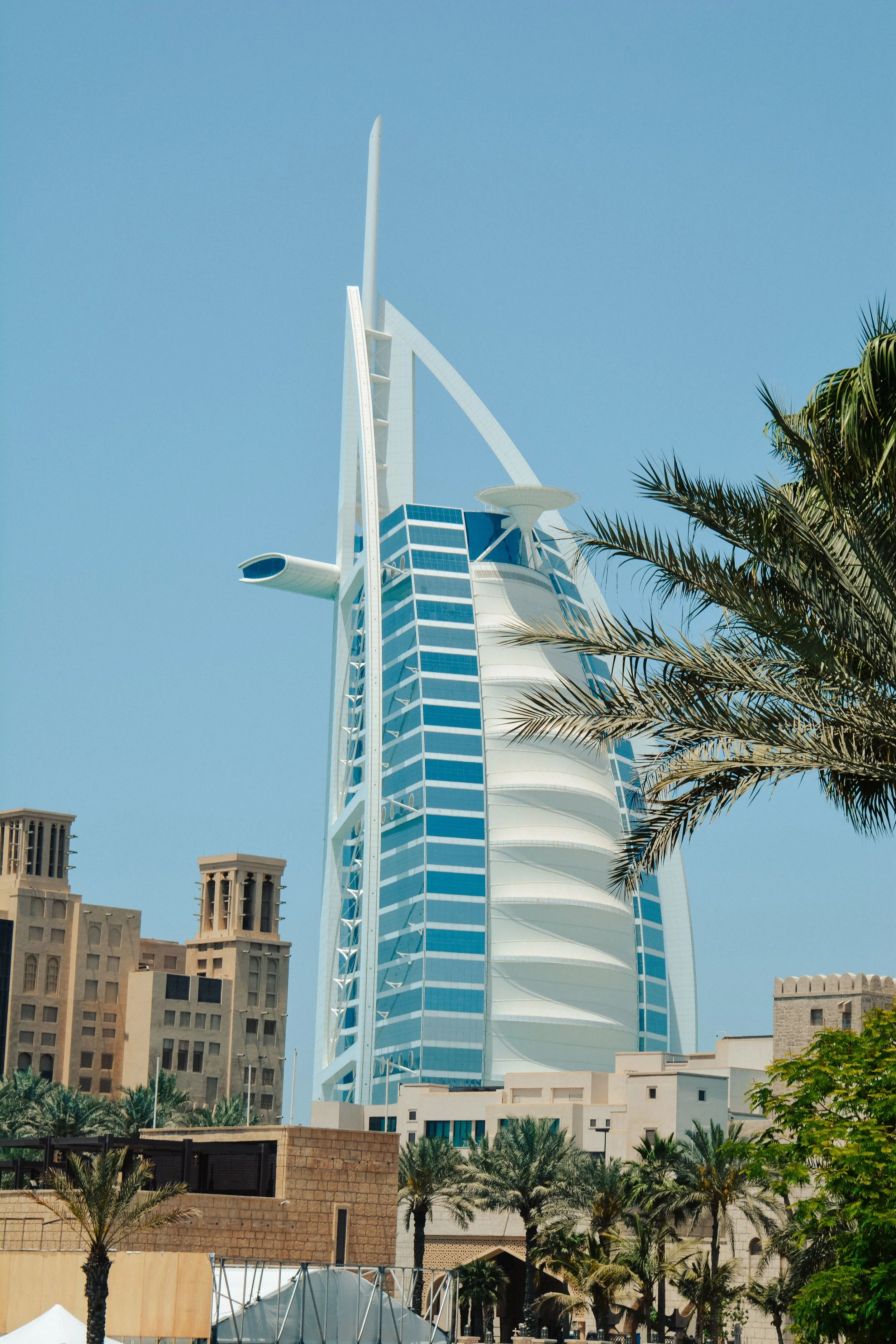 Tall, modern skyscraper with a distinctive sail-like design in Dubai, surrounded by palm trees and older buildings in the foreground, under a clear blue sky.