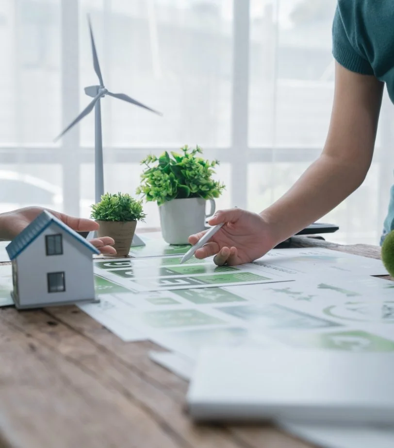 Twee mensen die een ontwerp bespreken op een tafel met miniatuurhuisje, planten, een model windturbine, en papieren met ontwerpen.