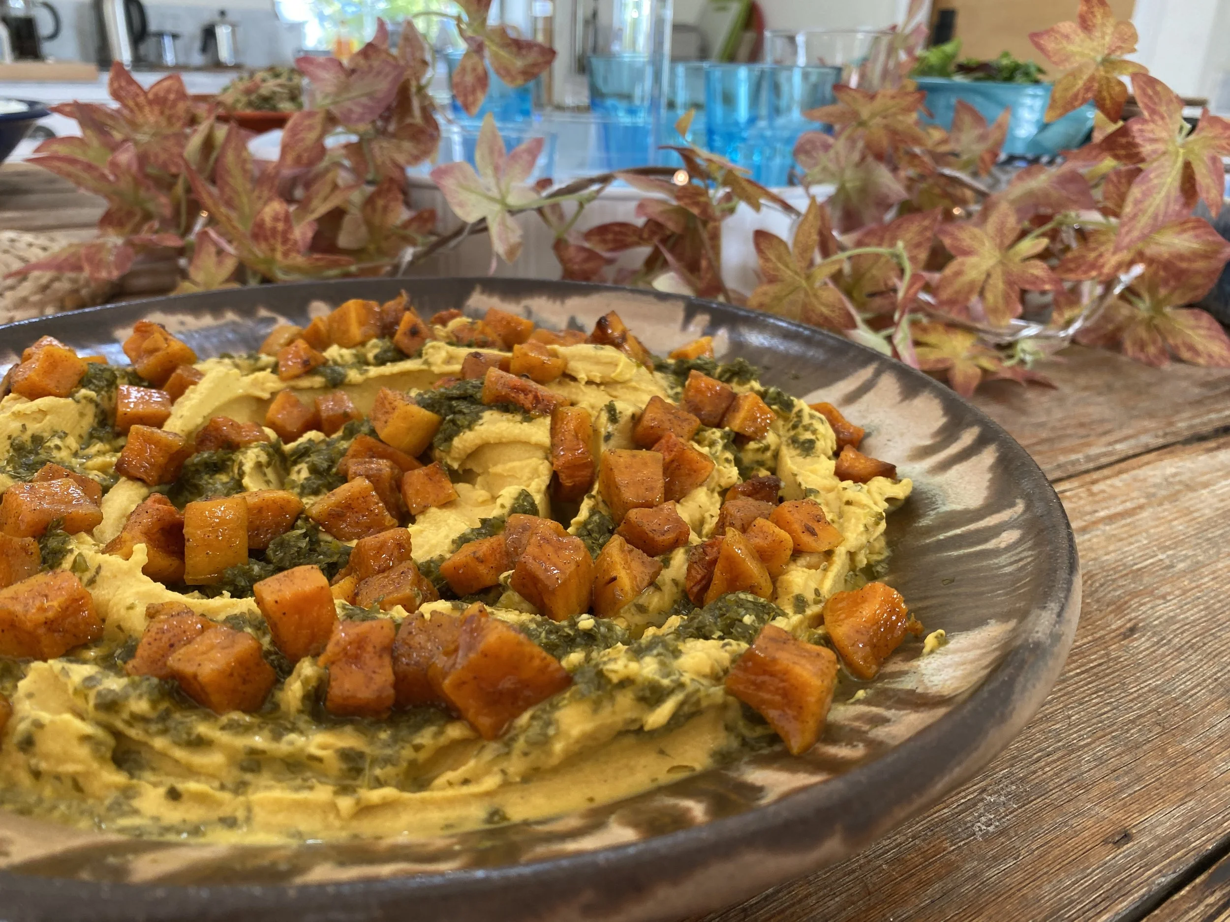 Close-up of a dish with scrambled eggs, diced sweet potatoes, and greens in a black bowl, with a background of colorful plants and glasses on a wooden table.