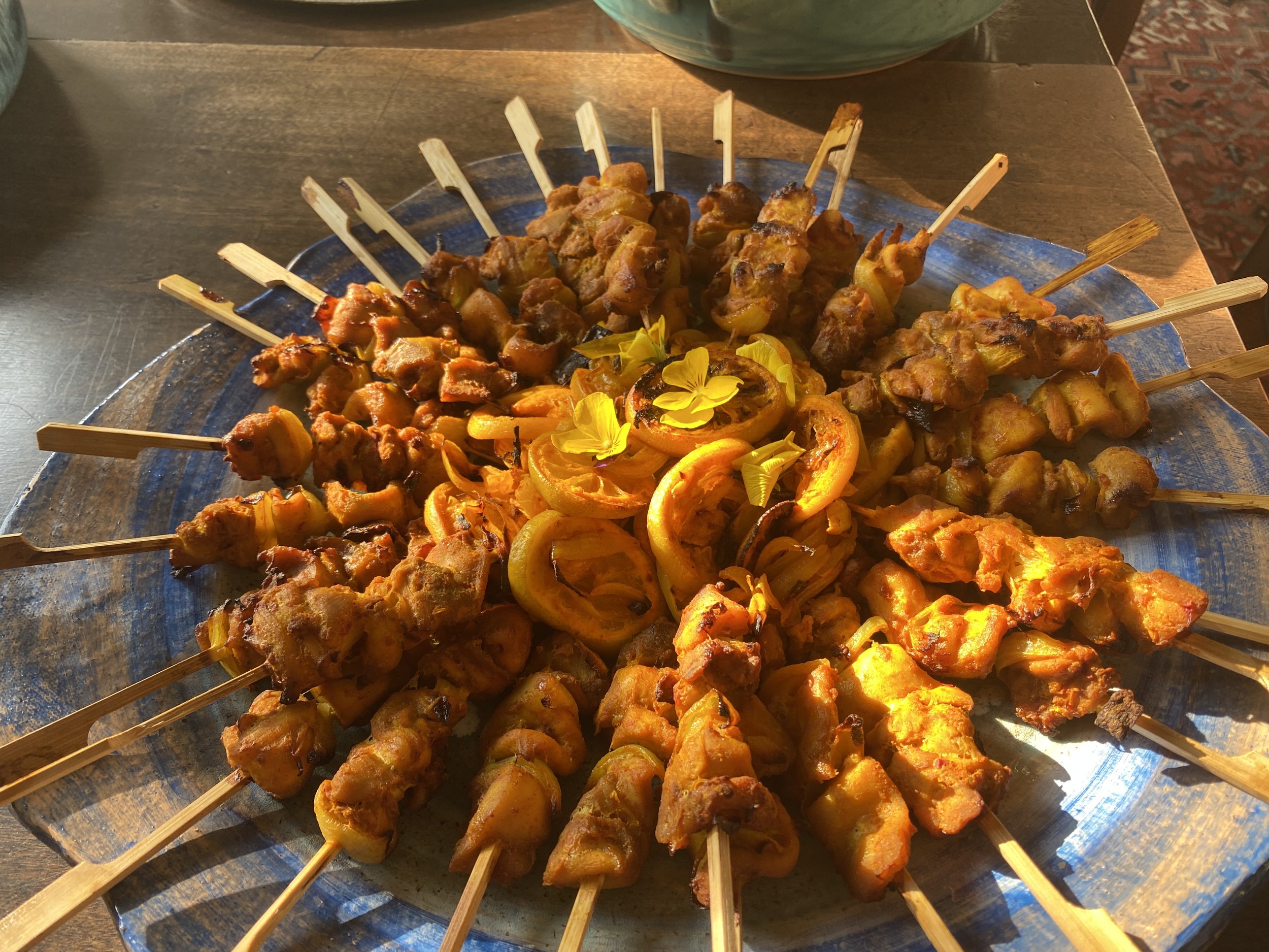 Skewers with grilled meat and vegetables arranged on a large round tray, decorated with yellow edible flowers.