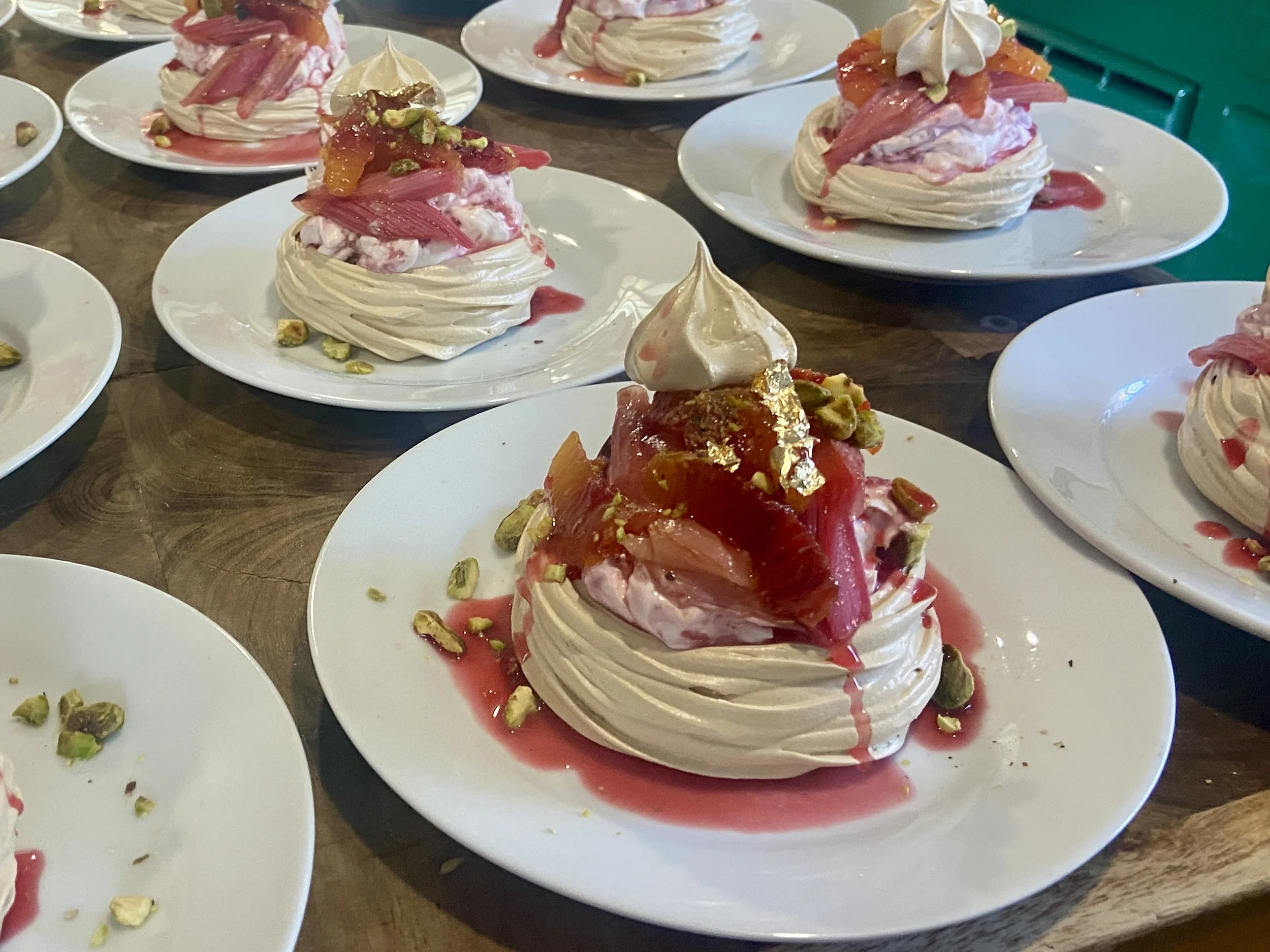 Several plates of pavlova decorated with pink fruit, fruit sauce, nuts, and cream on a wooden table.