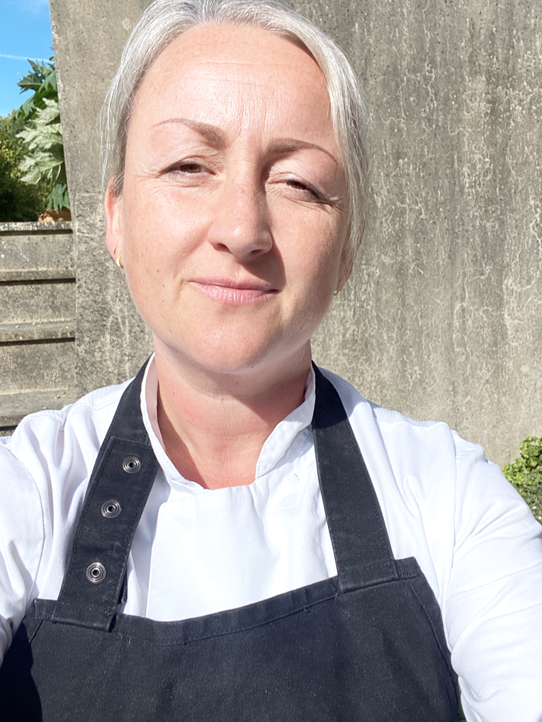 A woman with short blonde hair wearing a white chef's uniform and black apron, standing outside on a sunny day.