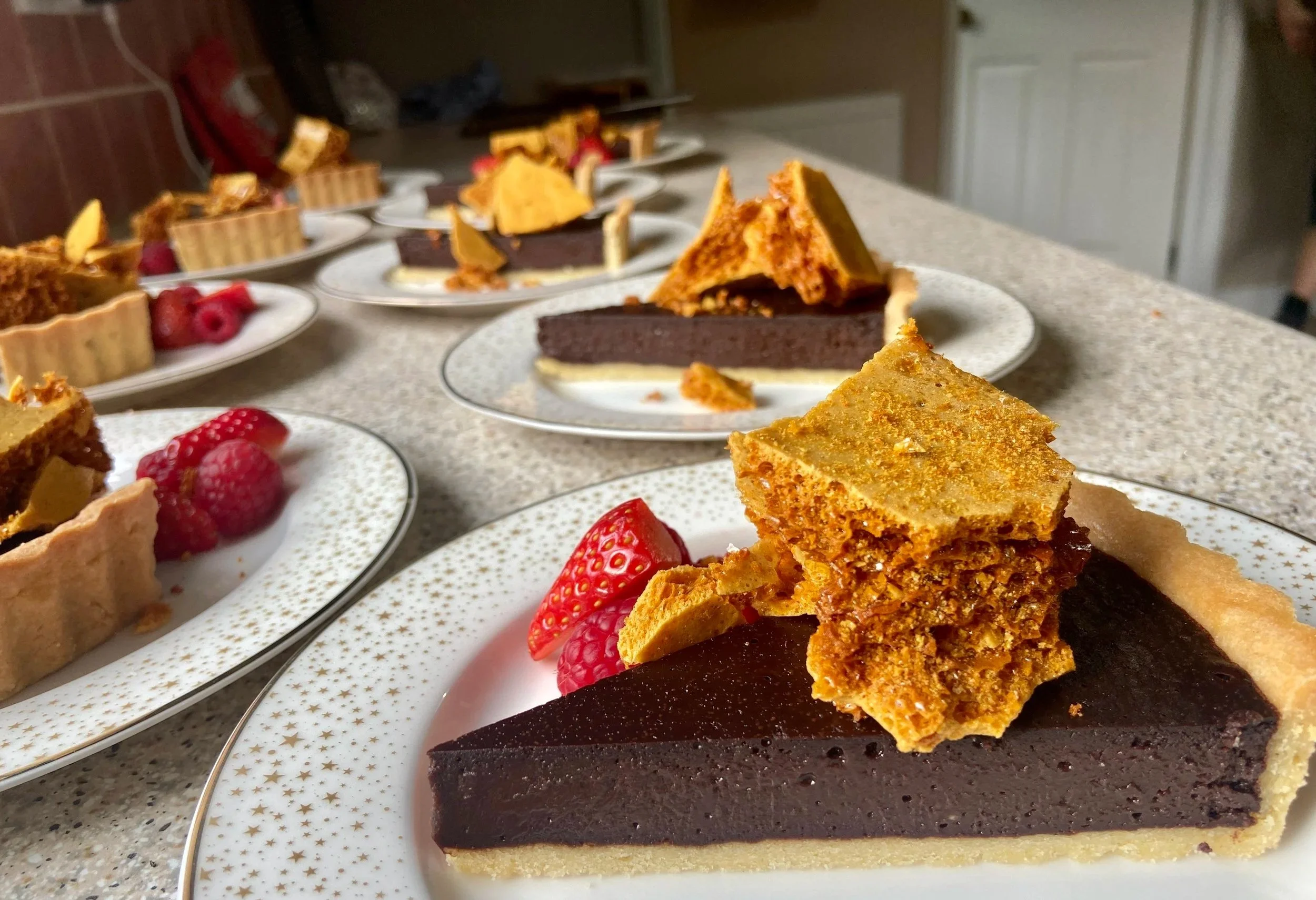 Multiple plates of assorted desserts including slices of chocolate cake, fruit tart, and strawberry-topped tart on a countertop.