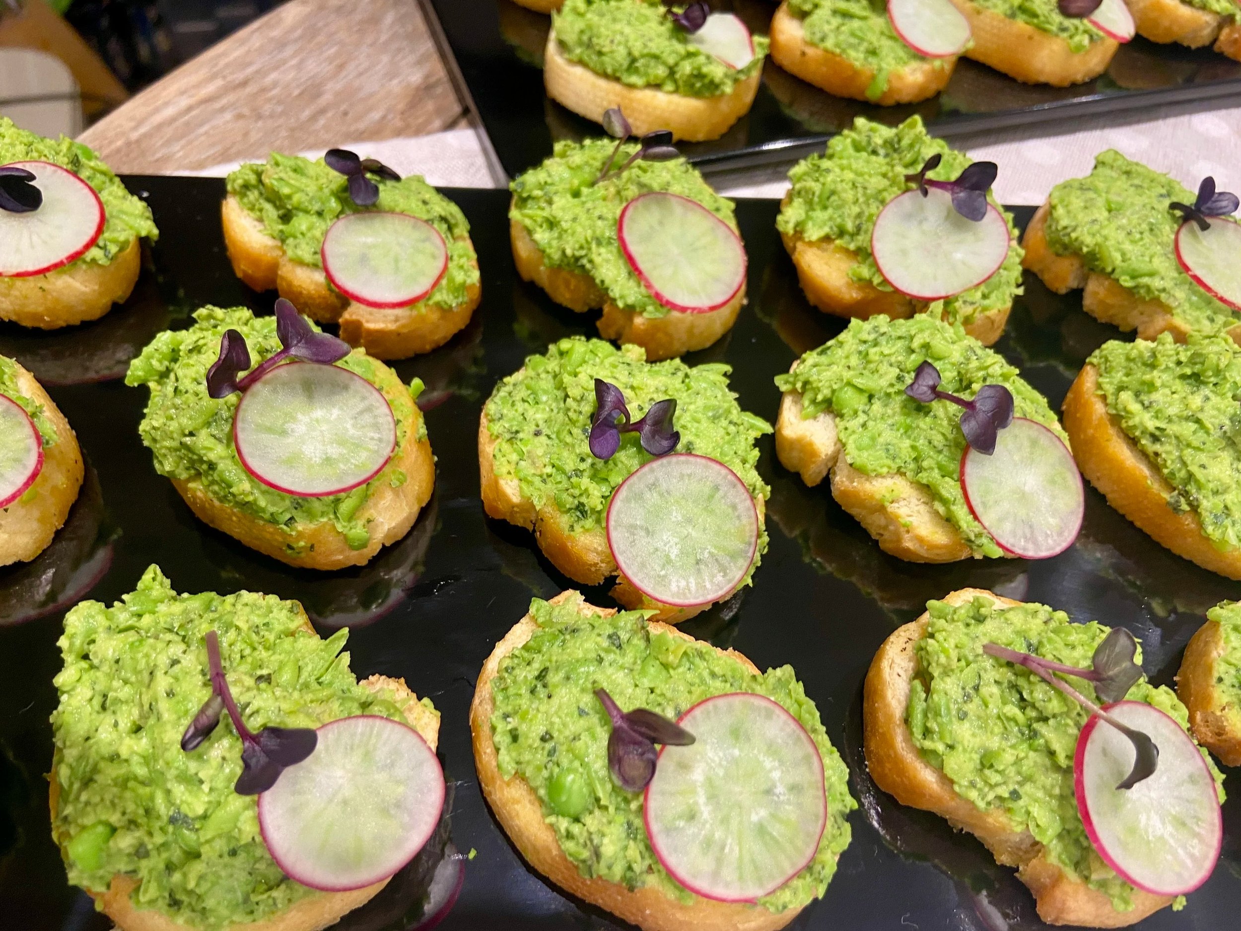 Close-up view of toasted bread slices topped with mashed avocado, garnished with sliced radishes and microgreens, arranged on a black platter.