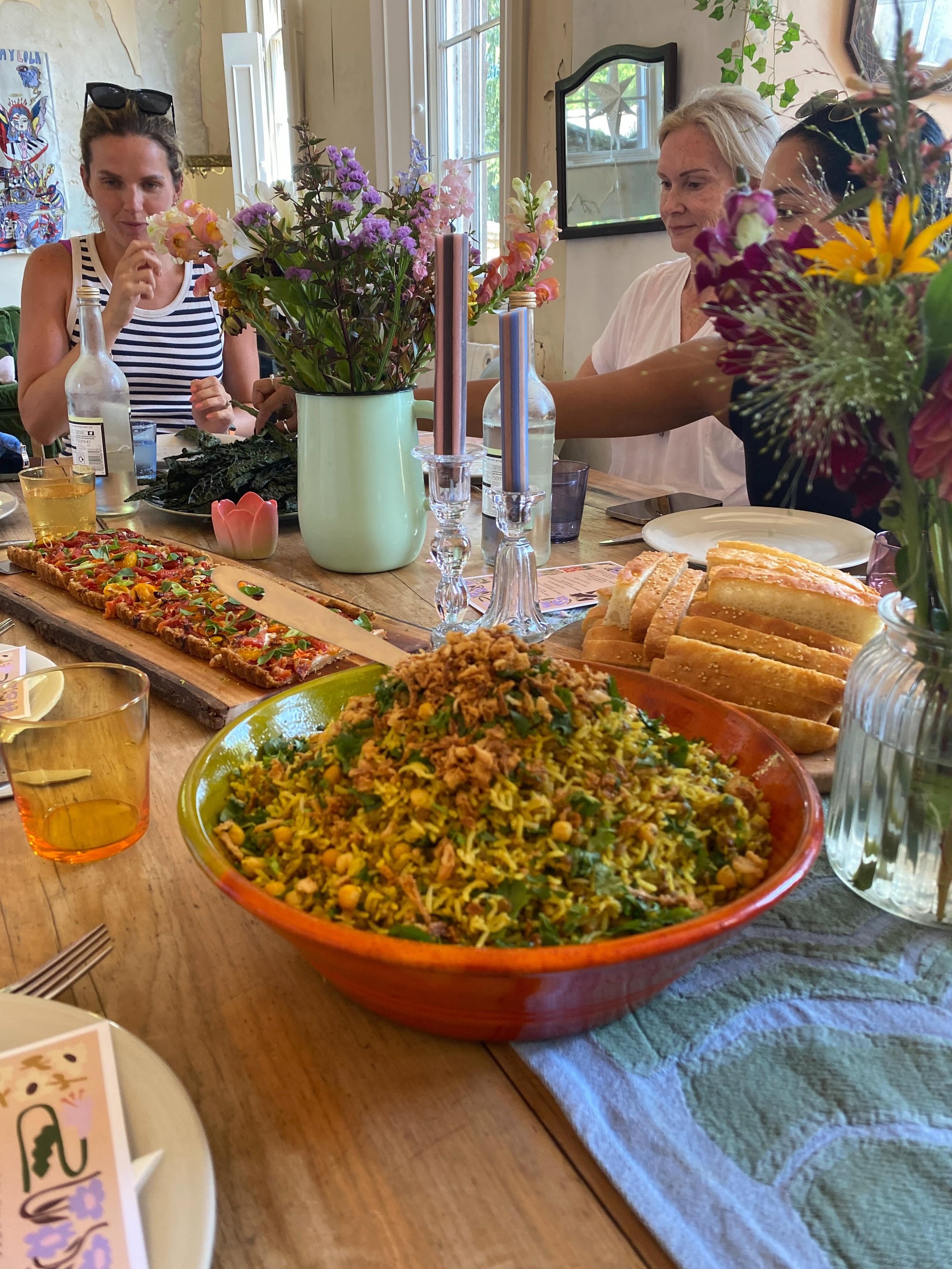 A table with a variety of colorful dishes including a large bowl of rice and vegetables, a flatbread pizza, sliced bread, and beverages, decorated with flowers and candles in a cozy, well-lit room.