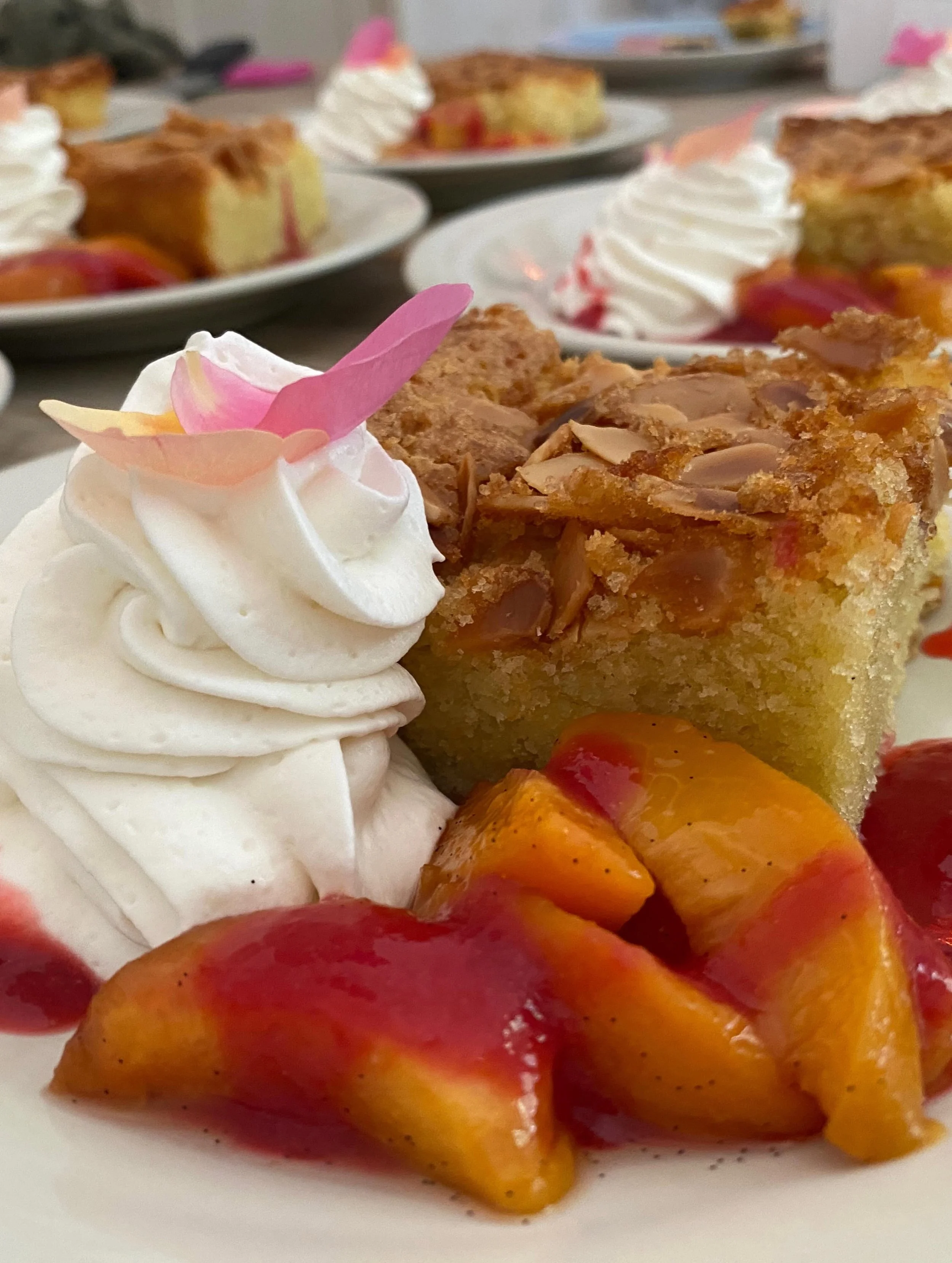 Close-up of a plate with a slice of peach cobbler topped with whipped cream and pink petals, with additional peach cobbler and whipped cream on plates in the background.