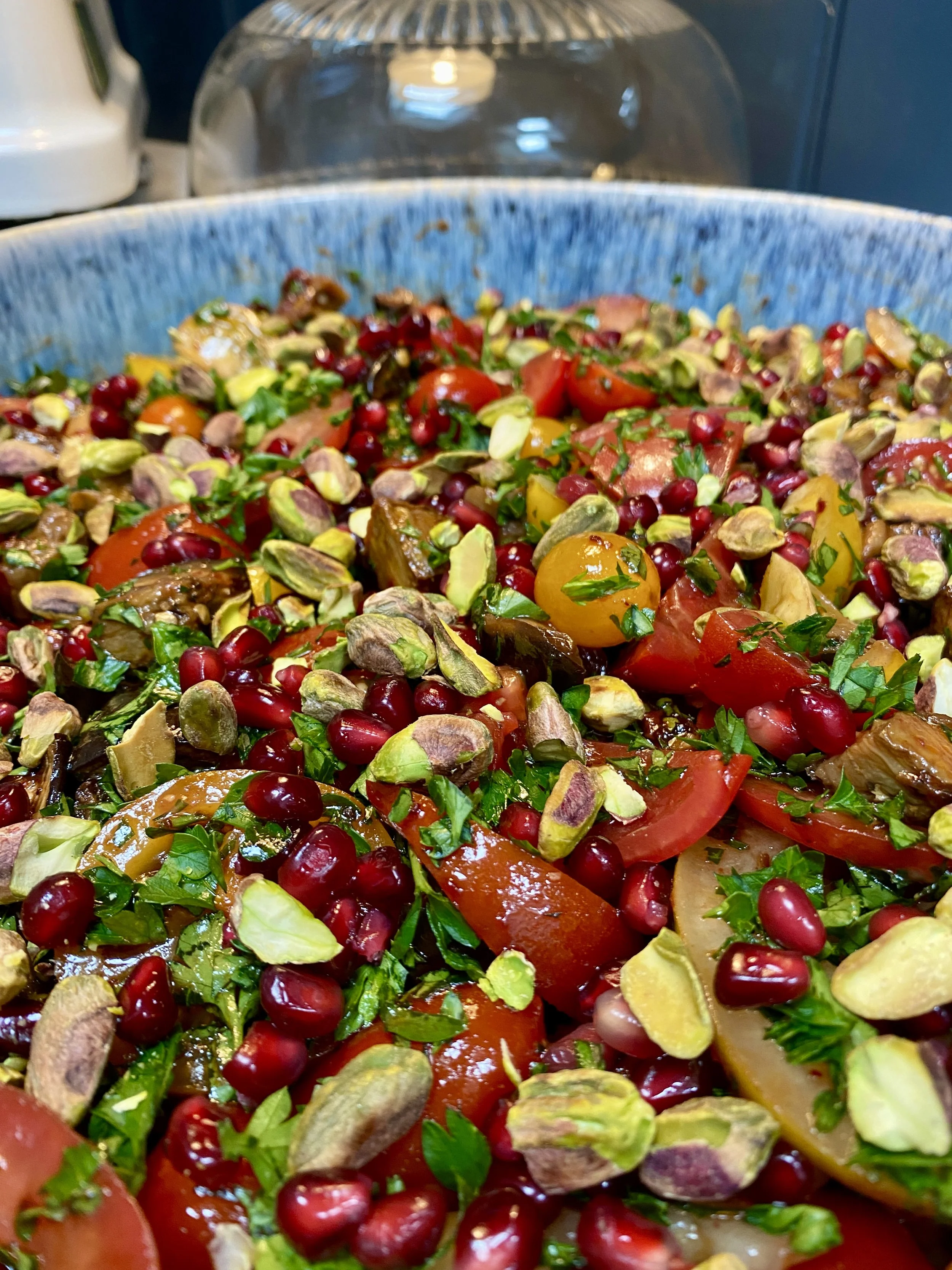 Close-up of a mixed salad in a blue bowl with cherry tomatoes, pomegranate seeds, chopped greens, and pistach nuts.