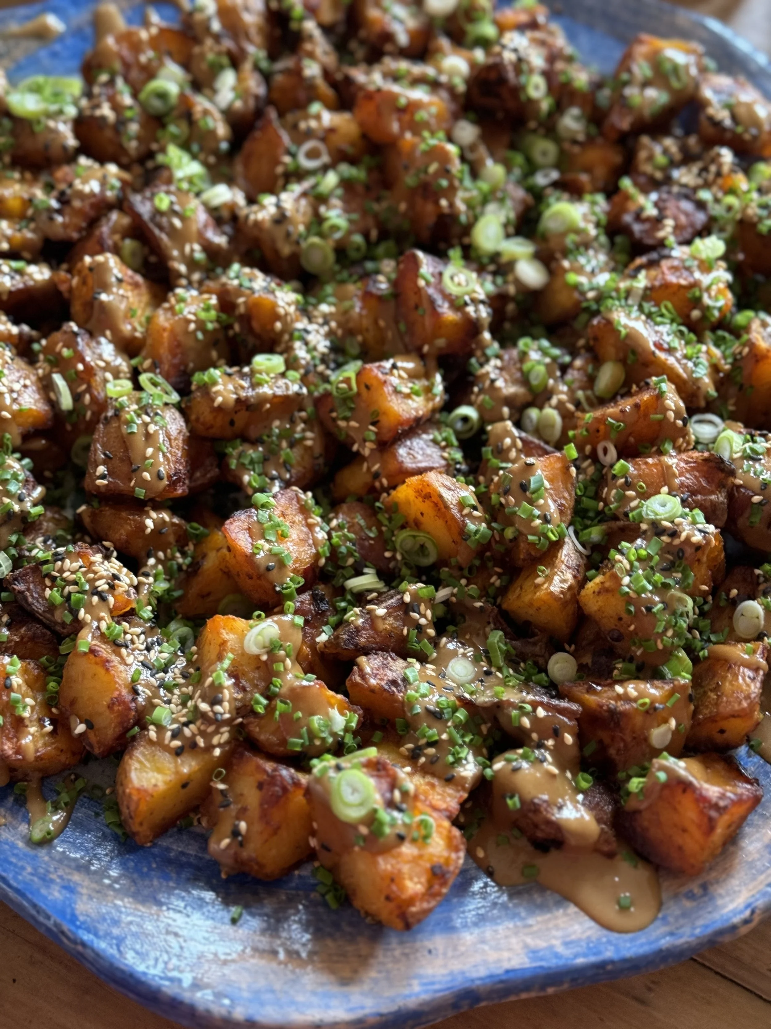 A plate of roasted sweet potatoes garnished with chopped green onions, sesame seeds, and drizzled with a sauce.