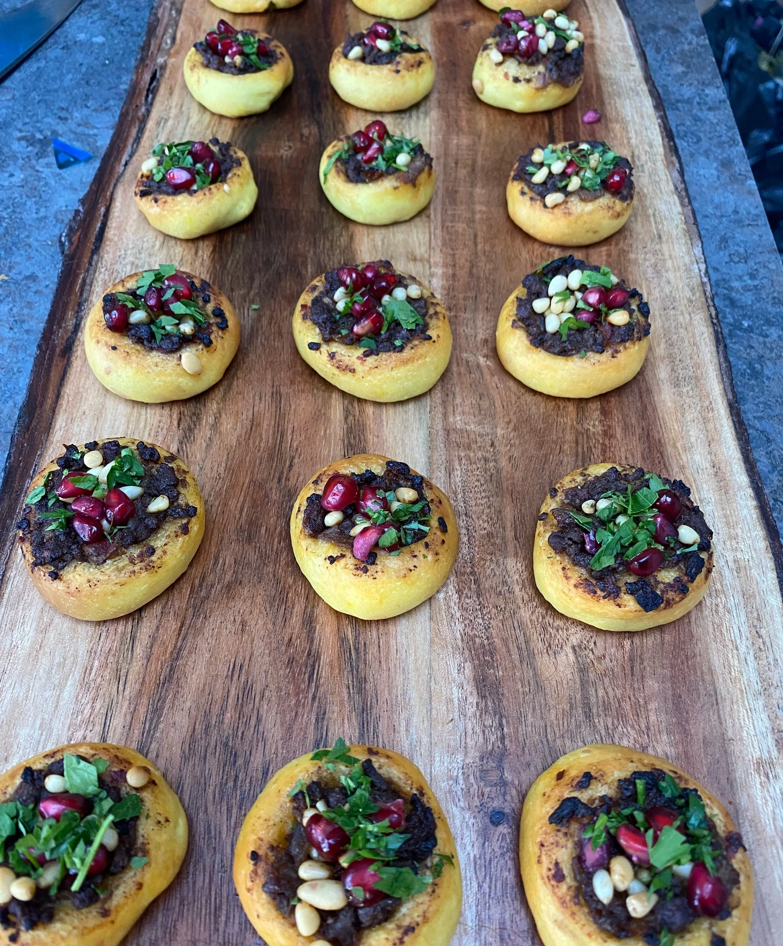 Close-up of grilled yellow squash halves topped with ground meat, pomegranate seeds, pine nuts, and chopped herbs on a wooden serving board.
